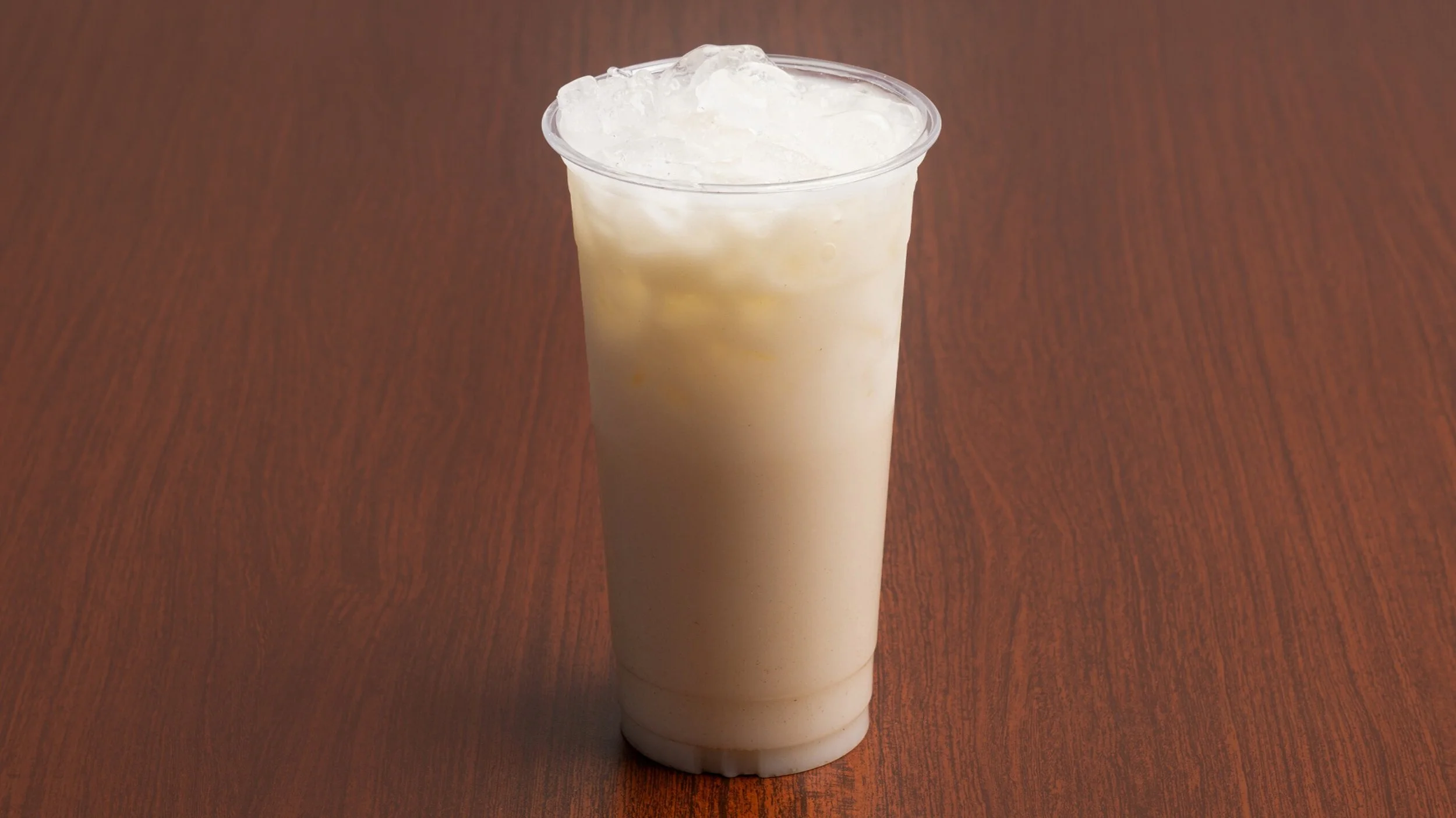 A clear plastic cup filled with vanilla milkshake topped with whipped cream, placed on a wooden surface.