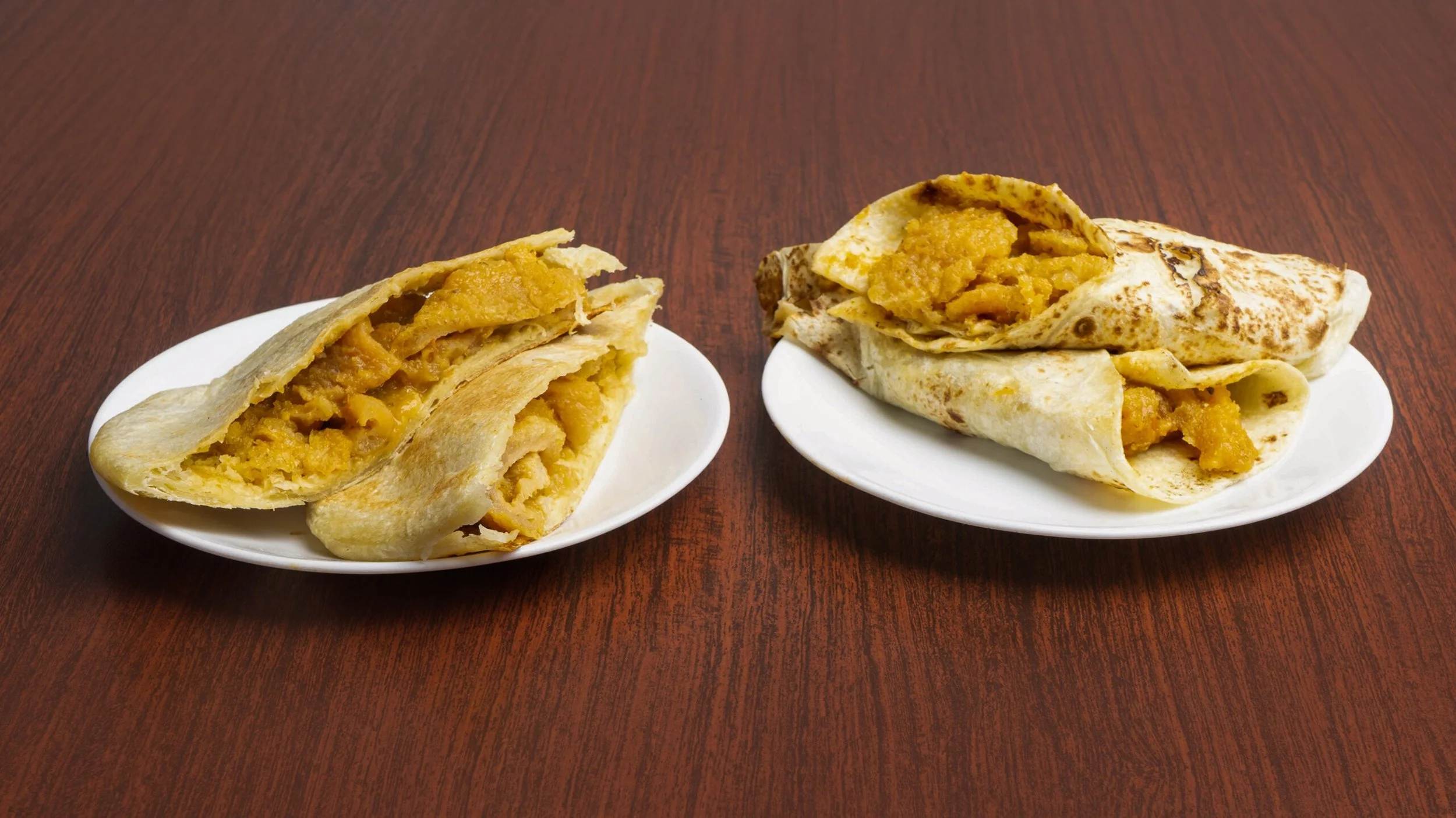 Two plates of Indian stuffed flatbreads, one cut in half, showing a bright yellow potato filling inside, placed on a wooden table.