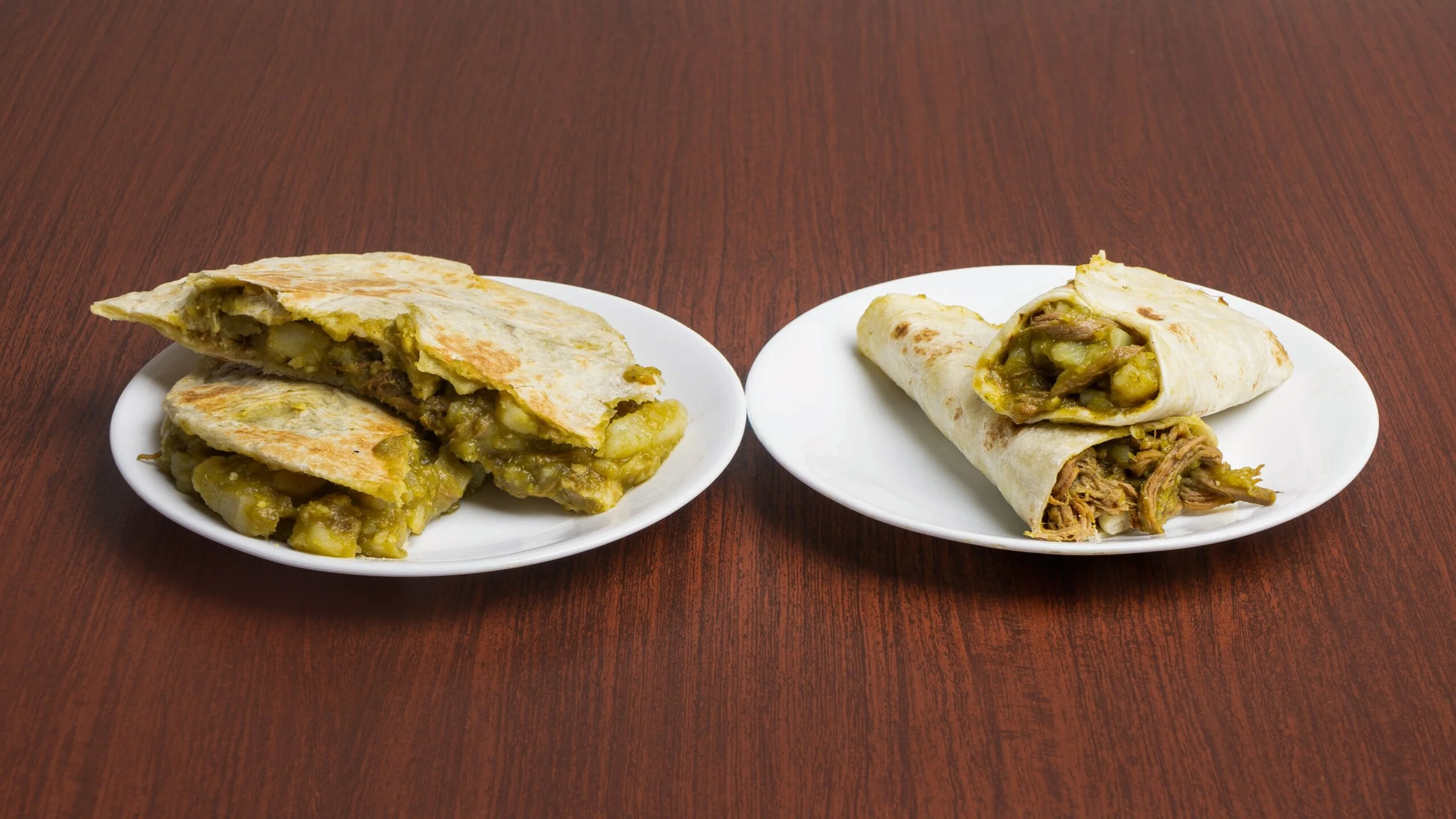Two white plates with Mexican tacos on a wooden table. The taco on the left is cut open, showing potatoes, meat, and sauce. The taco on the right is also cut open, displaying shredded meat, potatoes, and sauce.