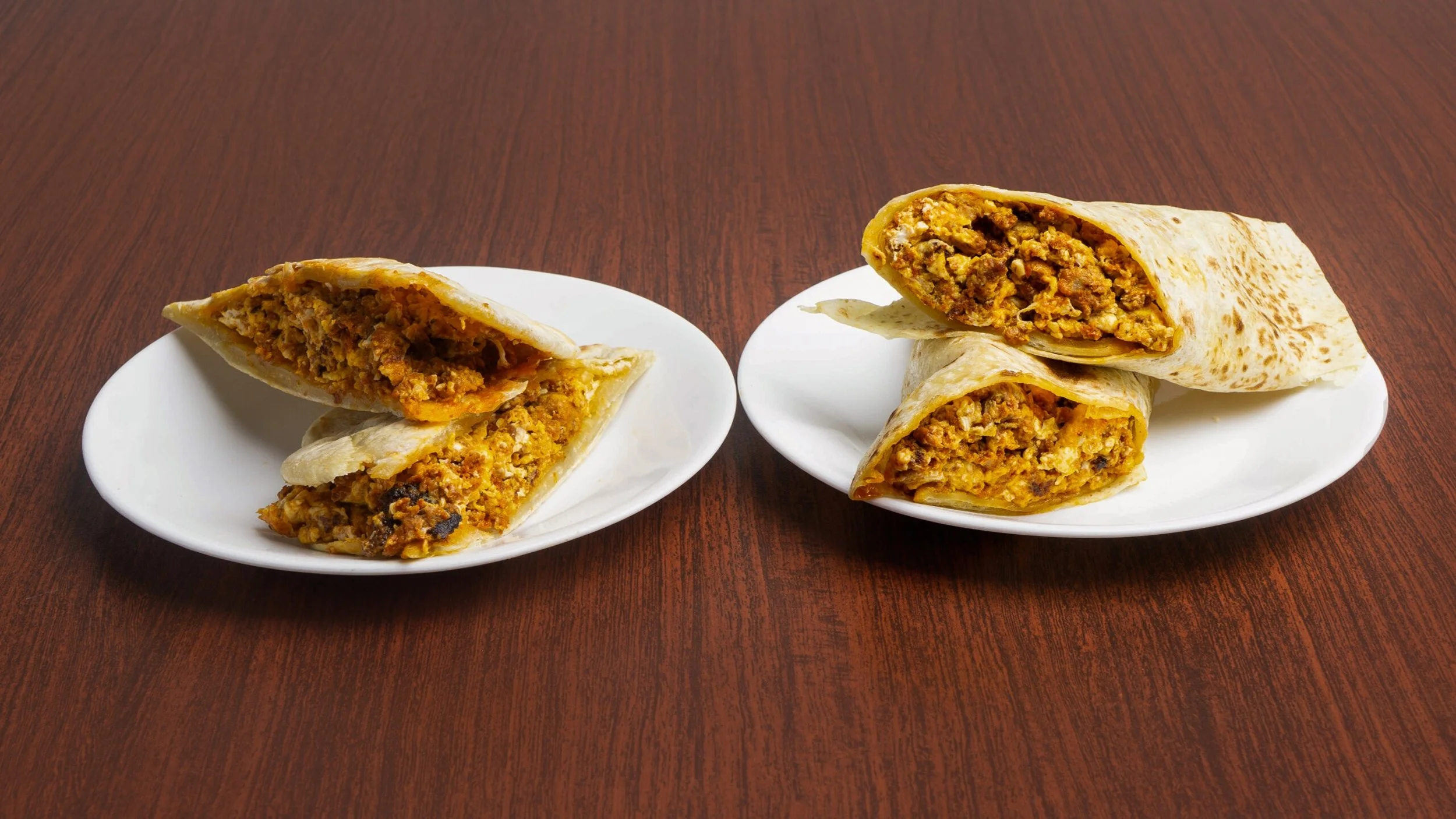 Three breakfast burritos on two white plates placed on a wooden table, each cut in half revealing scrambled eggs, sausage, and melted cheese inside a flour tortilla.