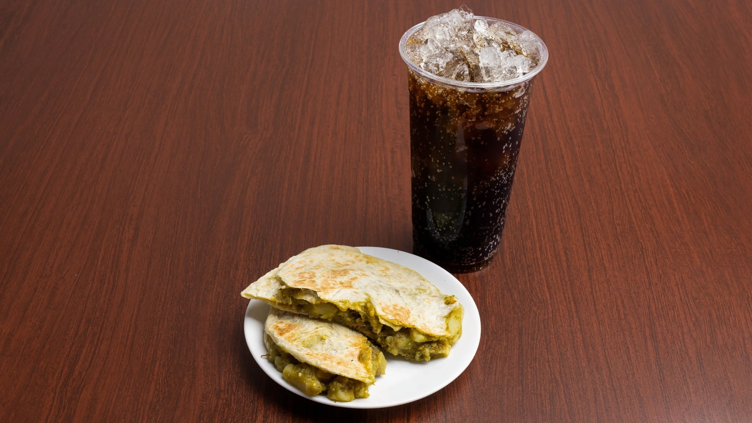 A plate with a cut quesadilla filled with vegetables next to a tall glass of soda with ice on a wooden table.