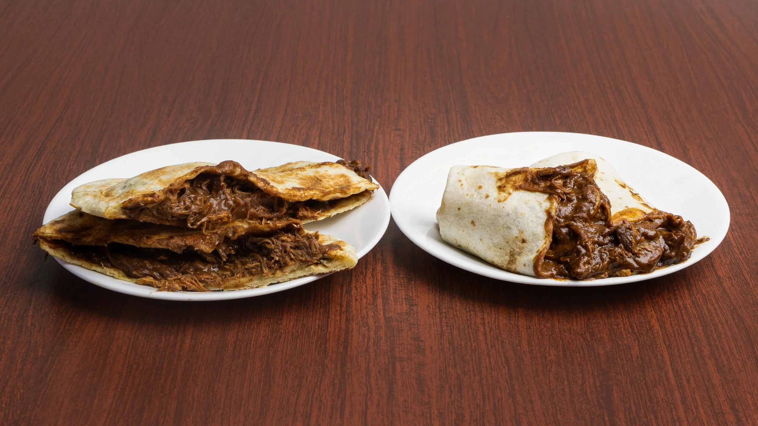 Two white plates with beef burritos on a wooden table, one cut open showing shredded beef and sauce inside, the other wrapped.