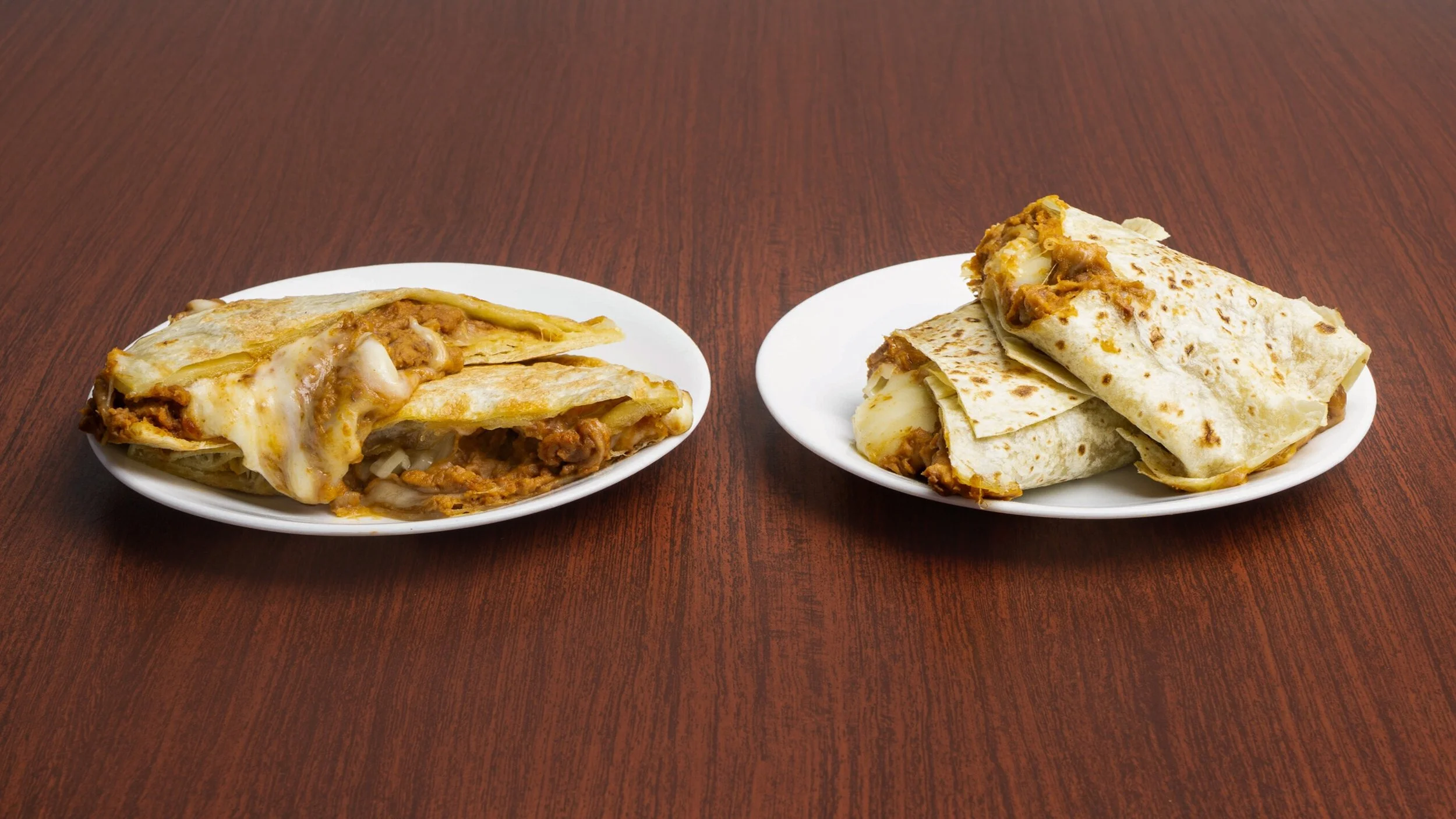 Two plates of Mexican-style burritos on a wooden table. The burritos are wrapped in flour tortillas, with one opened to show filling of meat, cheese, and sauce.