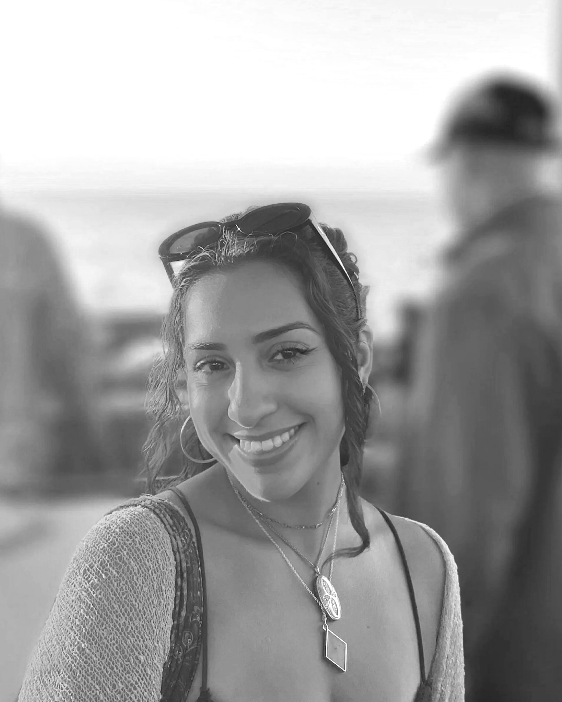 Smiling woman with wavy hair wearing sunglasses on her head, layered necklaces, hoop earrings, and a light-colored lace top, outdoors with a blurred background.