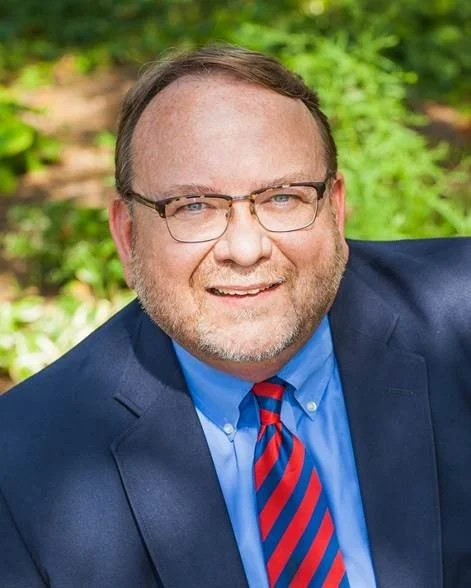 A middle-aged man with glasses, a beard, wearing a dark blue suit, light blue shirt, and a red and blue striped tie, smiling outdoors with green foliage in the background.