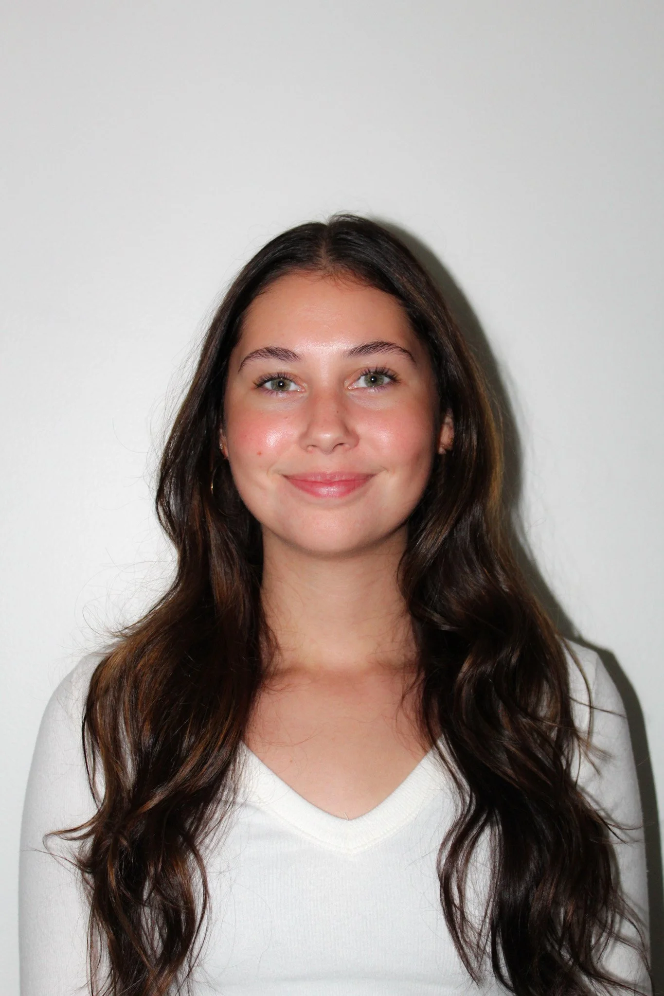 A young woman with long, wavy brown hair, light skin, and green eyes smiling in front of a plain white wall.