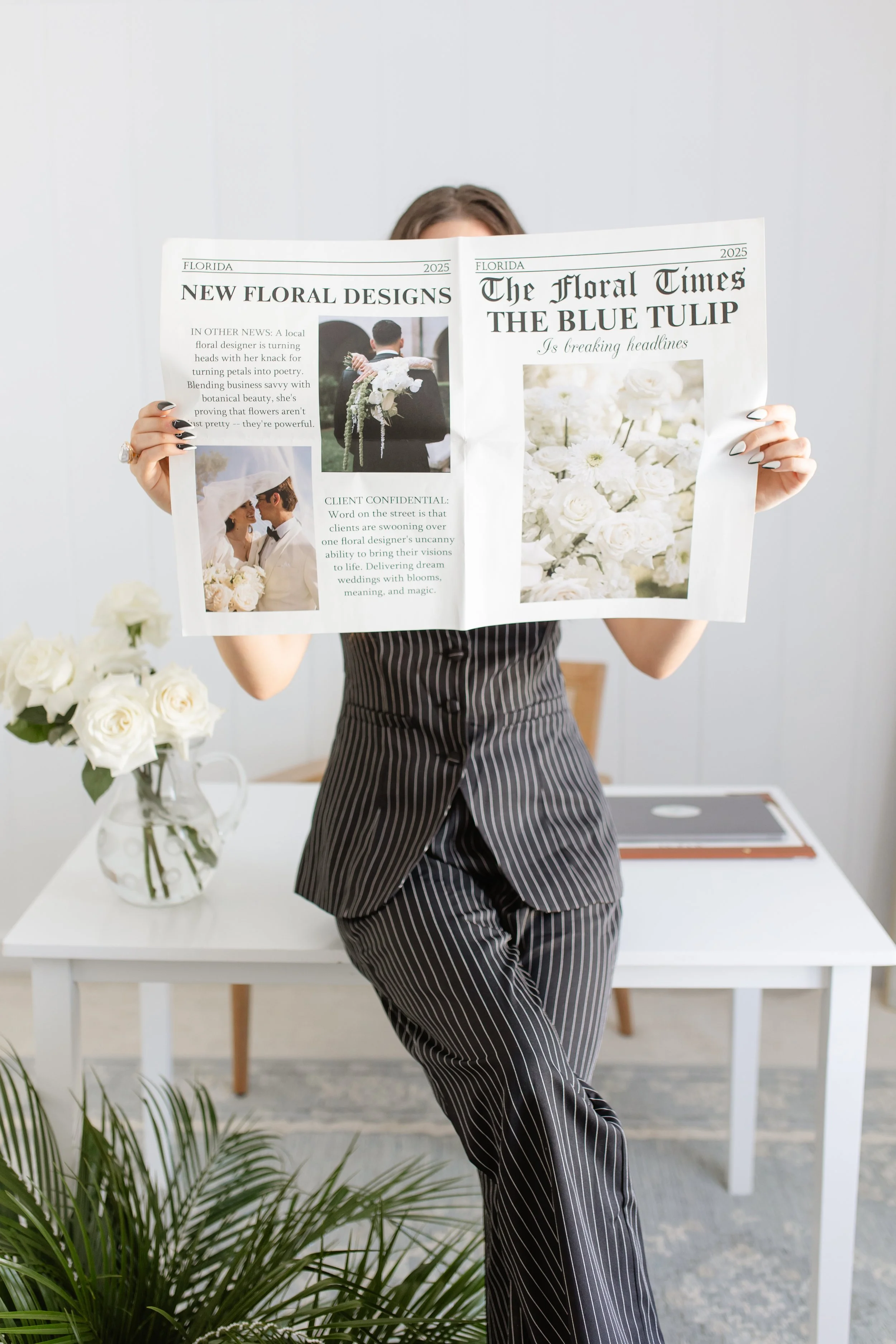 A woman in a striped dress is holding a newspaper with floral headlines in a bright room with a white table, a vase of white roses, and green plants. Professional wedding florist head shots.