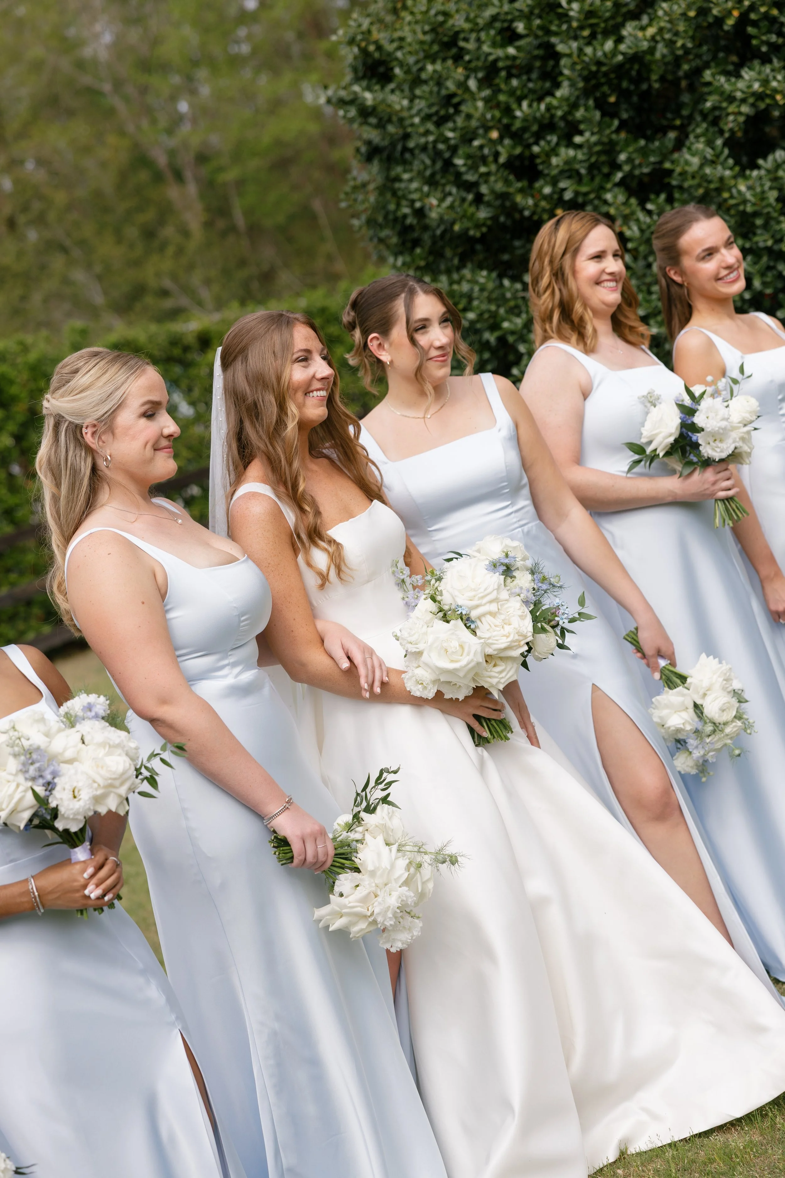 Group of bridesmaids wearing white dresses, holding bouquets of white flowers, standing outdoors with greenery in the background. Blue and white bridesmaid bouquets.