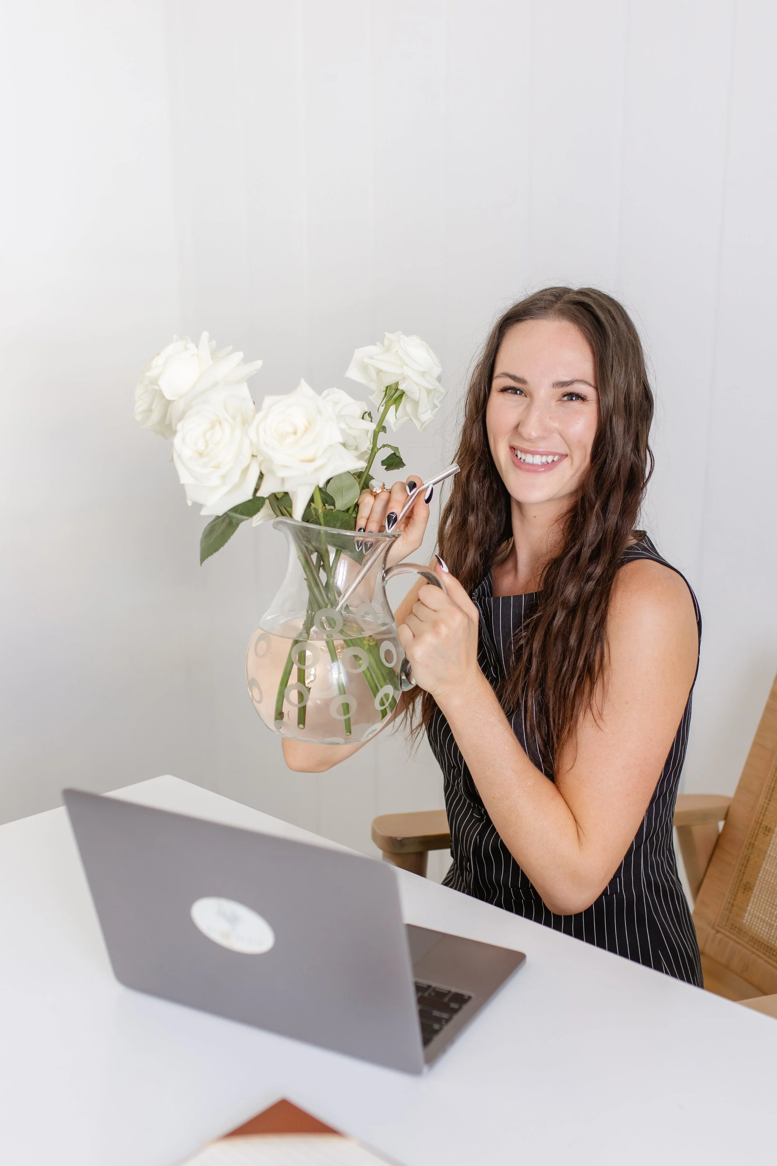 A woman with long brown hair smiling and holding a bouquet of white roses in a clear glass vase with a scoop handle, sitting at a white desk with a silver laptop in front of her. Wedding florist professional head shots.