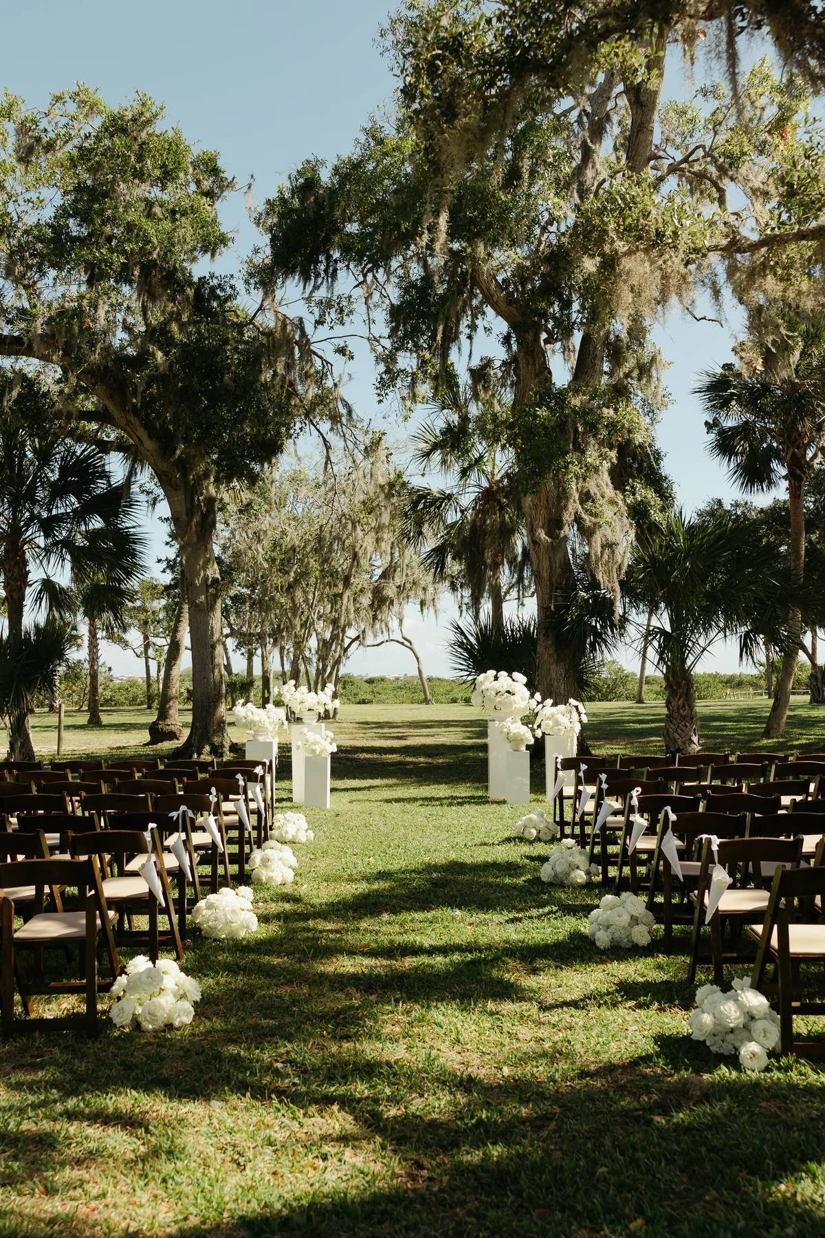An outdoor wedding venue decorated with white flower arrangements along the aisle, set under tall trees in a lush green outdoor setting.