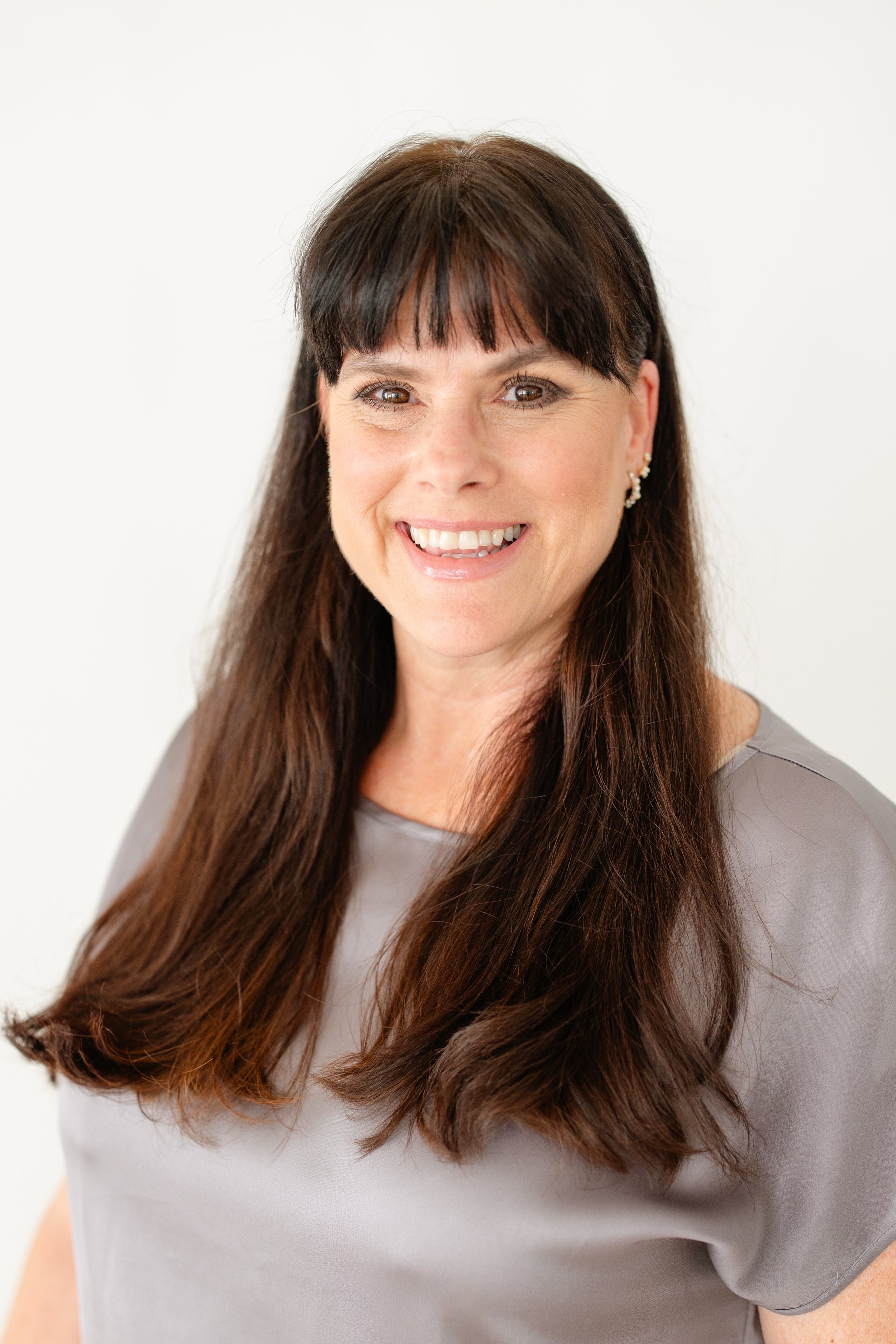 A woman with long brown hair, wearing earrings and a light gray top, smiling at the camera against a plain white background. Wedding florist professional head shots.
