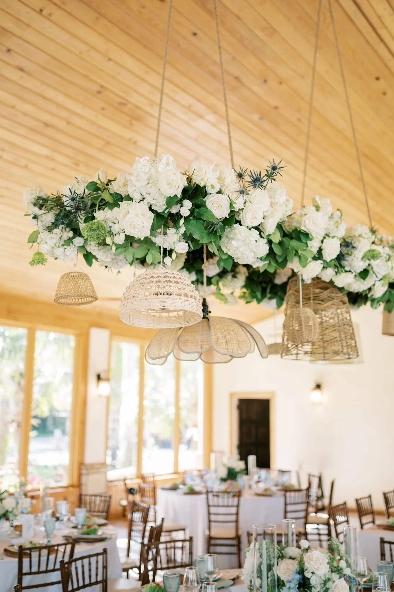 Indoor event space decorated with elegant floral arrangements featuring white roses and green foliage, hanging wicker and rattan lanterns, and set tables with glassware and place settings, with large windows and a wooden ceiling letting in natural light.