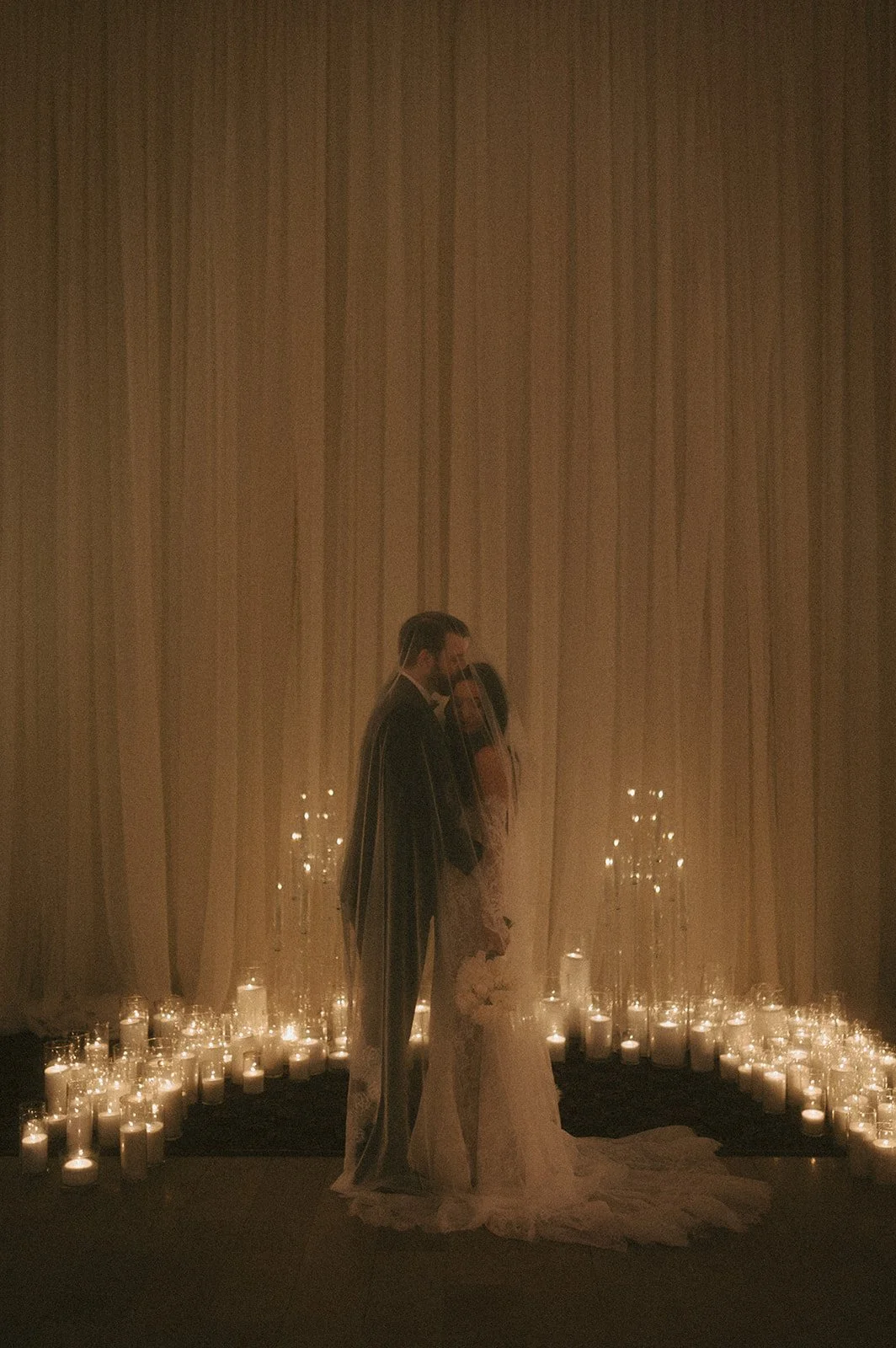 A bride and groom standing together surrounded by lit candles in glass jars, with a dimly lit backdrop. Crescent moon of candles surrounding couple. Candle Wedding.