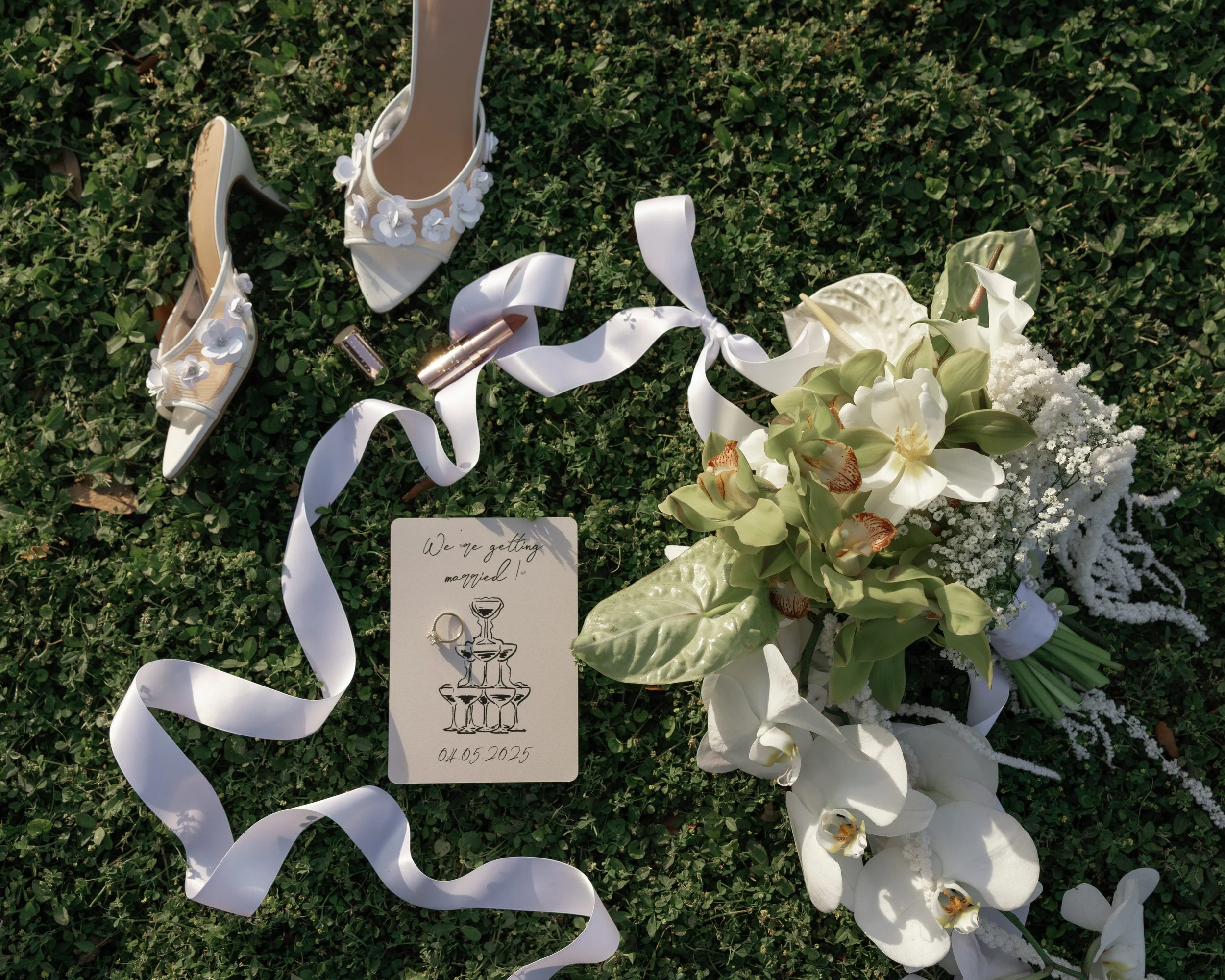 Wedding shoes, flowers, ribbons, and a sign reading 'We're getting married! 04.05.2025' on grass.