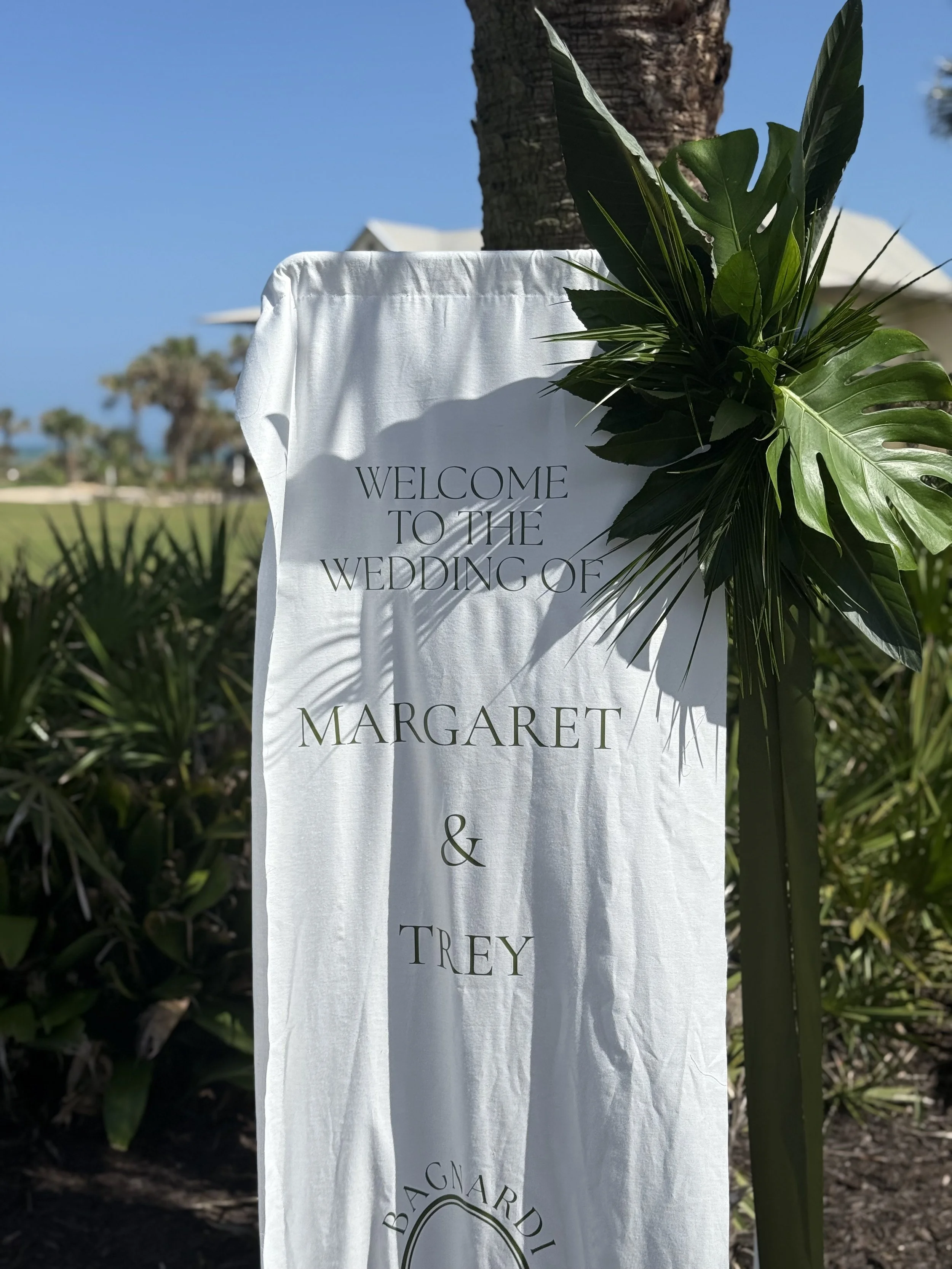 A white fabric banner with green leaves attached, reading 'Welcome to the wedding of Margaret & Trey', set outdoors with trees, blue sky, and a palm tree in the background. Tropical wedding palms and flowers for welcome sign.