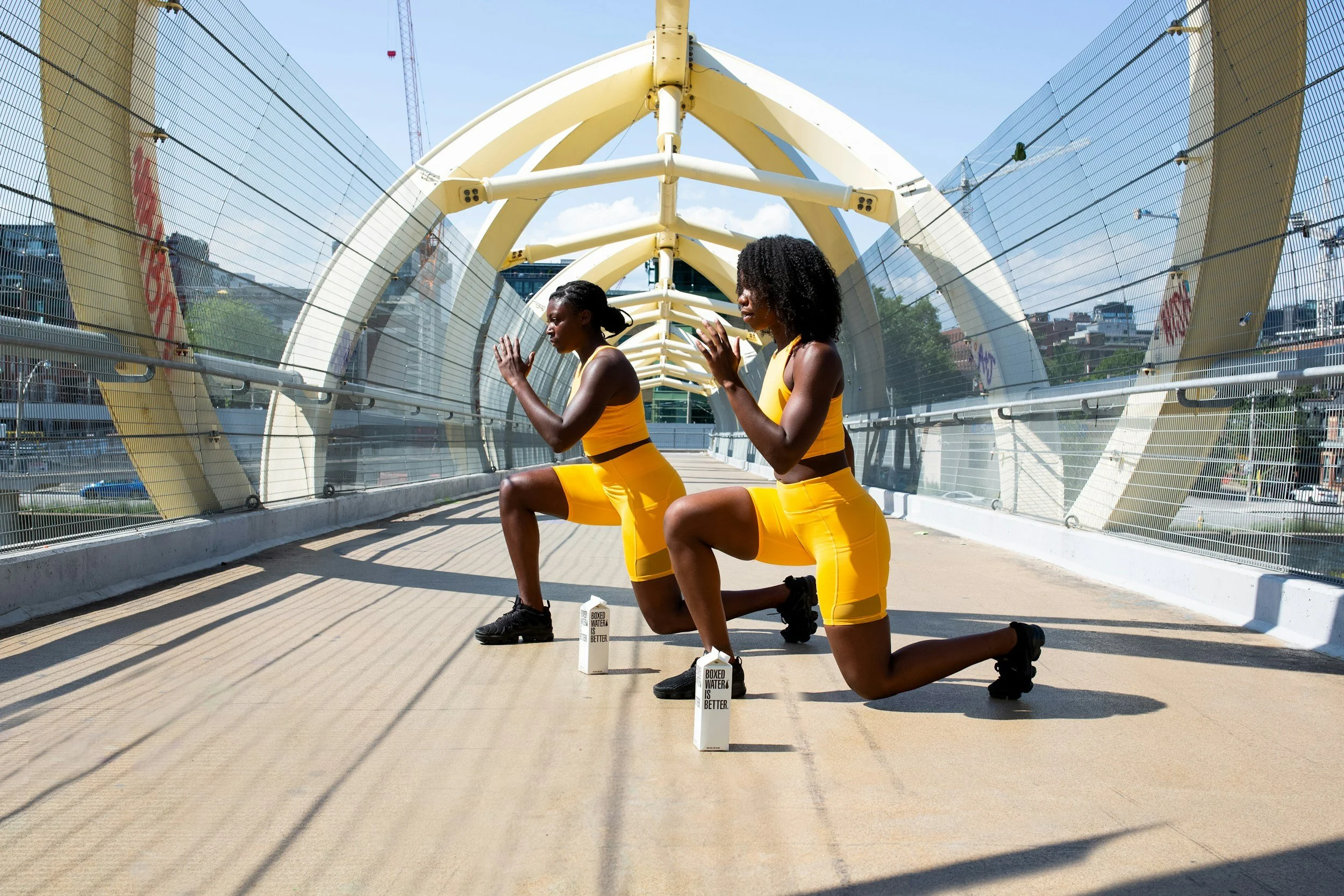 Two women wearing yellow workout outfits kneeling on one knee under a modern, arched pedestrian bridge, with their hands pressed together in prayer or meditation, and water bottles labeled 'Boxed Water Is Better' placed on the ground in front of them.