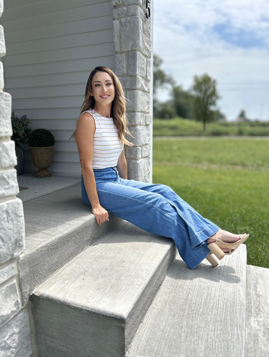 A woman with long wavy hair is sitting on concrete steps outside a house, smiling at the camera. She is wearing a white and beige striped sleeveless top, wide-leg blue jeans, and beige high-heeled sandals. The house has a stone and siding exterior, with a basket of greenery next to her. The background features a grassy area, trees, and a partly cloudy sky.