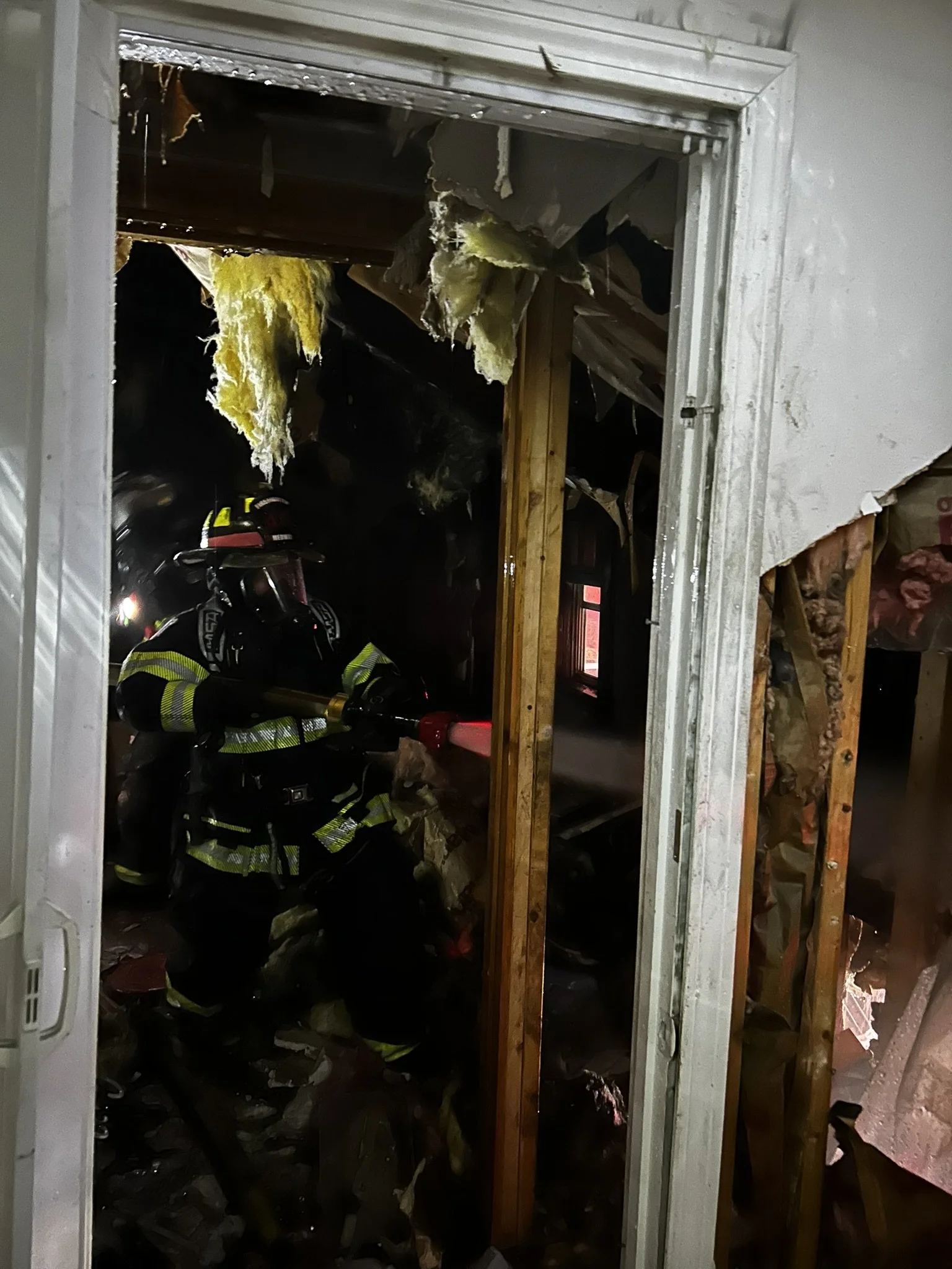 Firefighter in protective gear using a hose to spray water inside a room with extensive fire damage, charred debris, and damaged insulation visible.