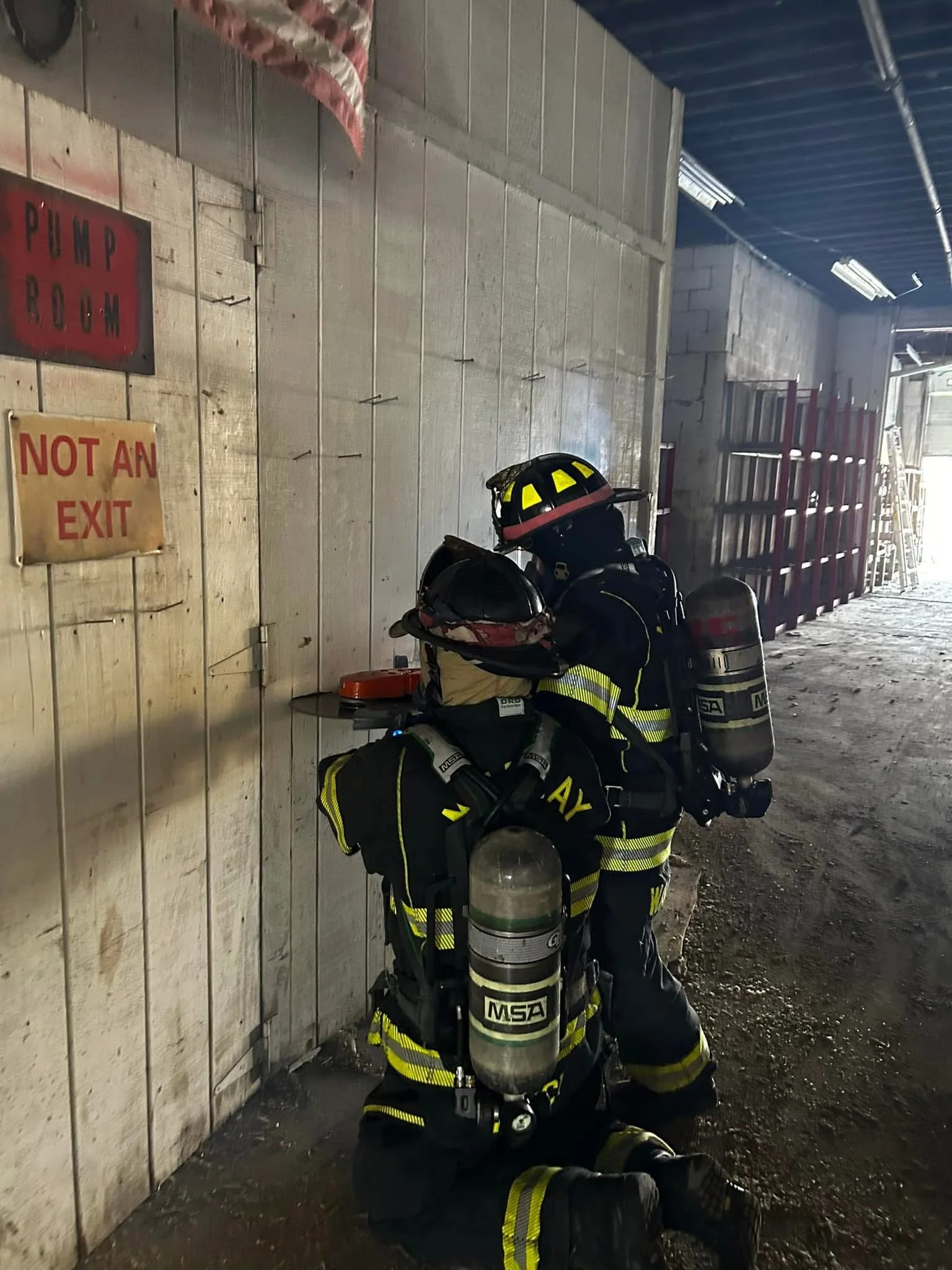 A firefighter wearing full gear, including a helmet with reflective stripes, kneeling on the ground and holding a helmet. The firefighter is near a wall with signs that read 'PUMP ROOM' and 'NOT AN EXIT'. The area appears to be an industrial or fire 