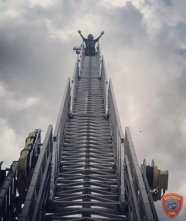A person sitting at the top of an emergency fire truck ladder with arms raised in the air, against a cloudy sky, with a Medway Fire Department logo visible in the bottom right corner.