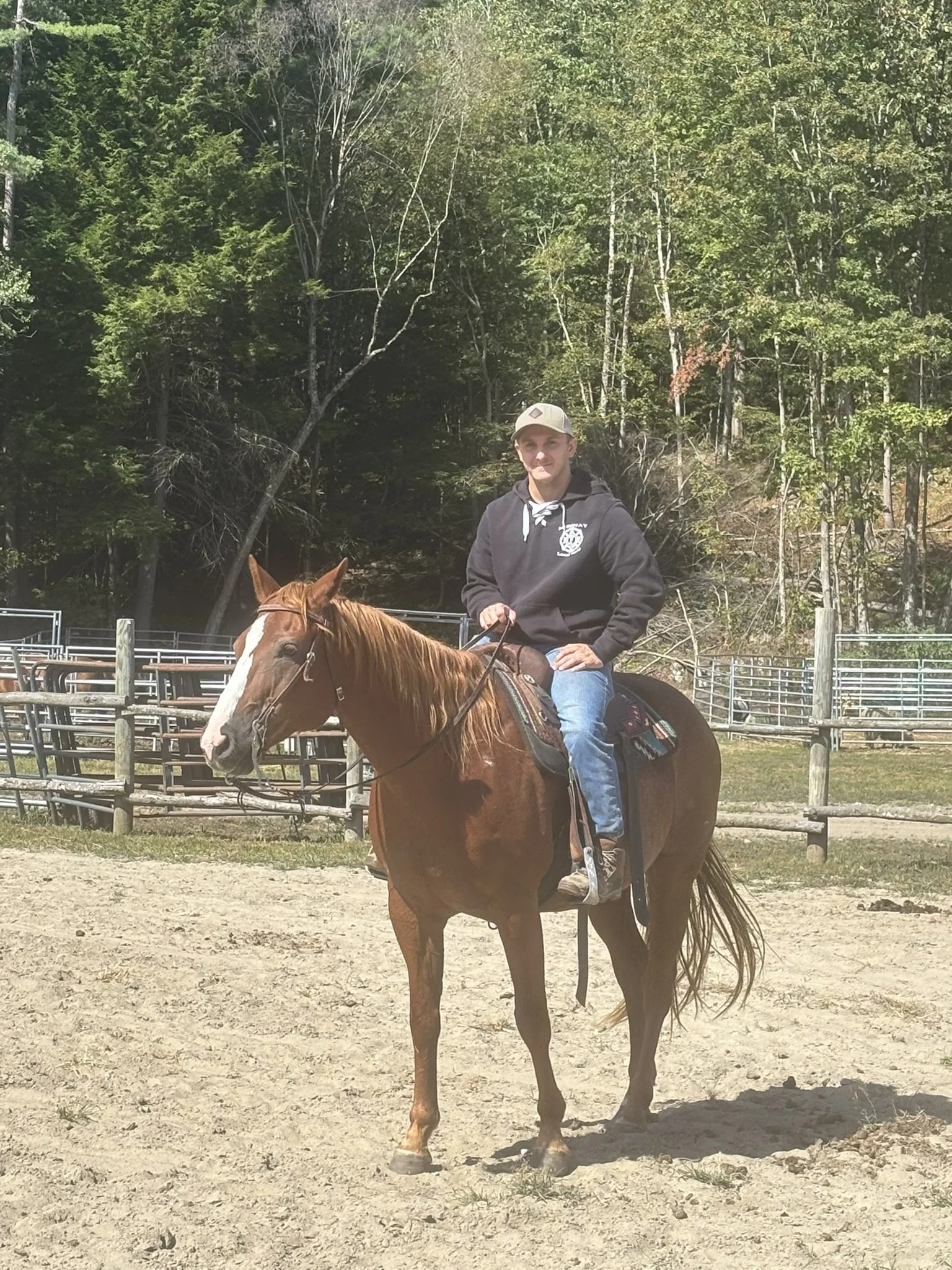 A young man wearing a gray baseball cap and a black hoodie riding a brown horse in an outdoor riding arena with trees and fencing in the background.
