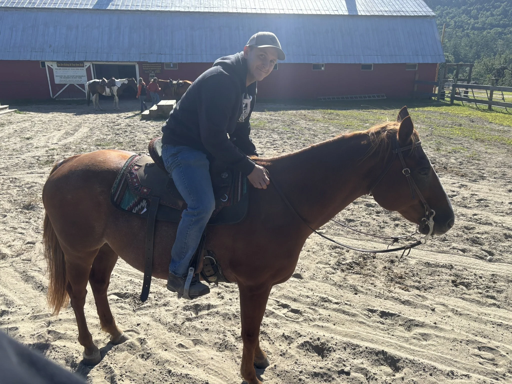 Person riding a chestnut horse on a sandy outdoor area near a barn with other people and horses in the background.