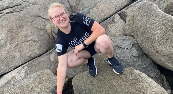 Woman with glasses crouching on large rocks outdoors, smiling at the camera