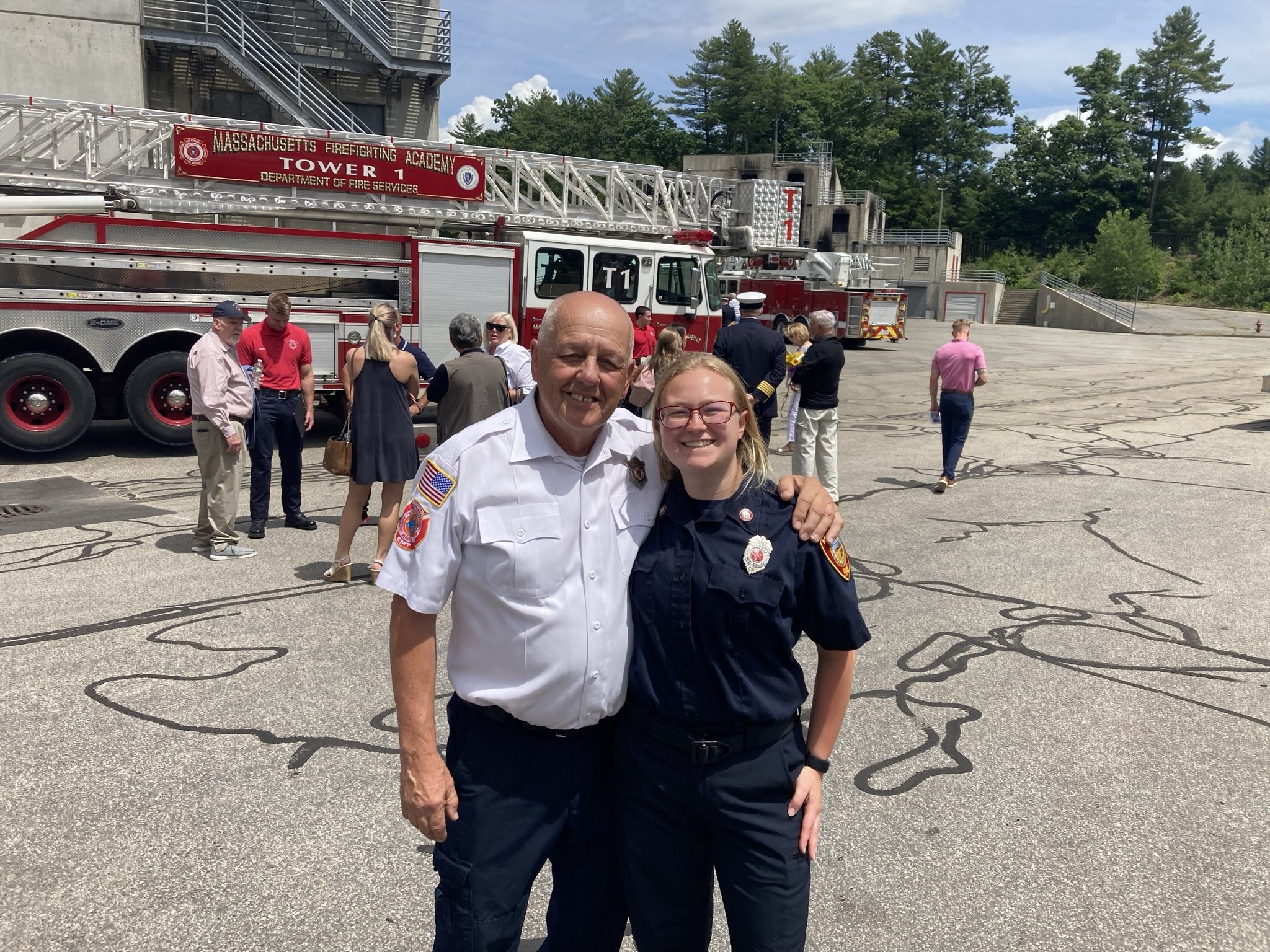 Two firefighters, a man and a woman, smiling and standing together in front of a fire truck, with a group of people and a fire station in the background.