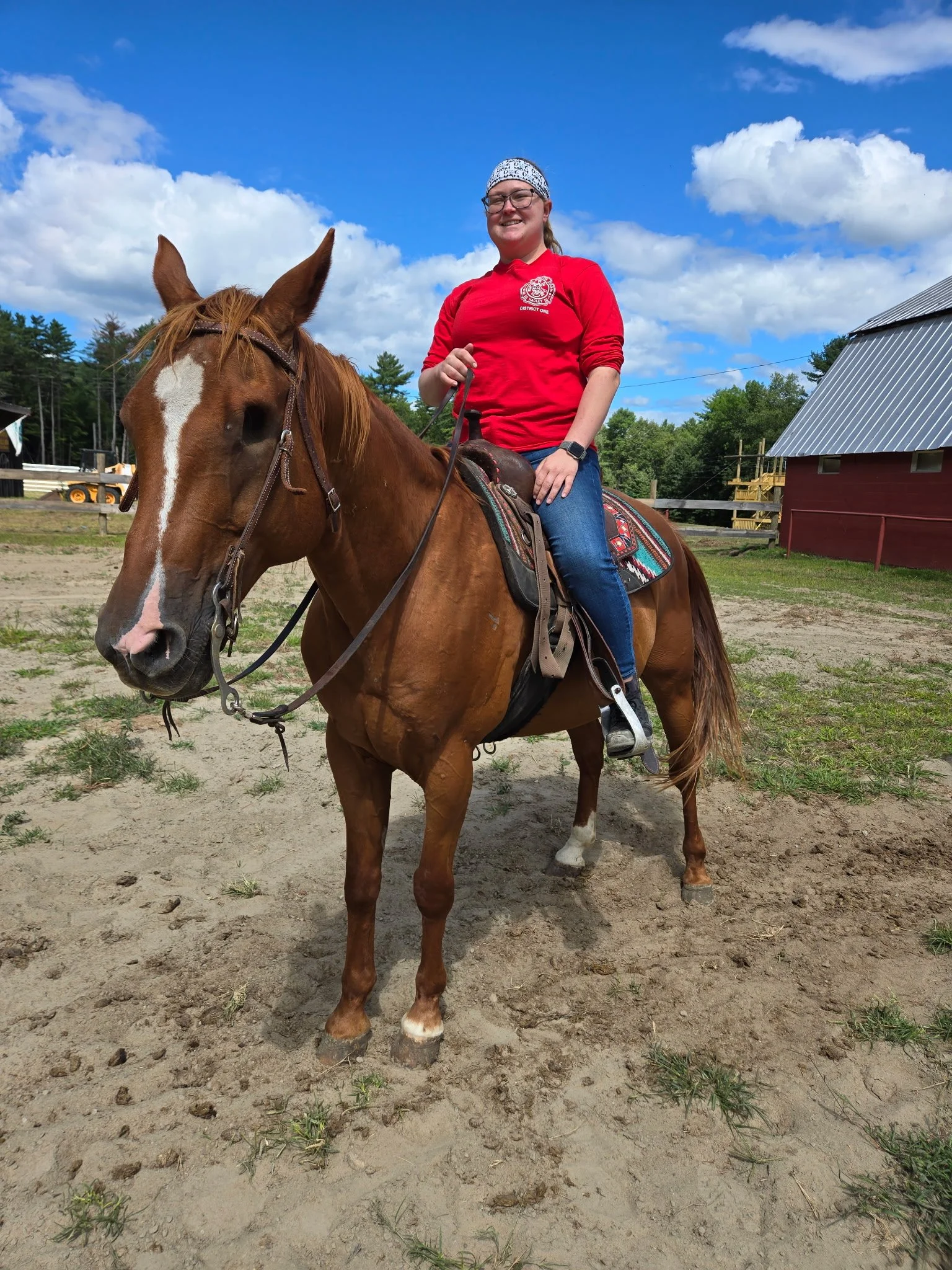A woman in a red shirt and jeans riding a brown/chestnut horse outdoors on a sunny day with a blue sky and white clouds, near a red barn and trees in the background.