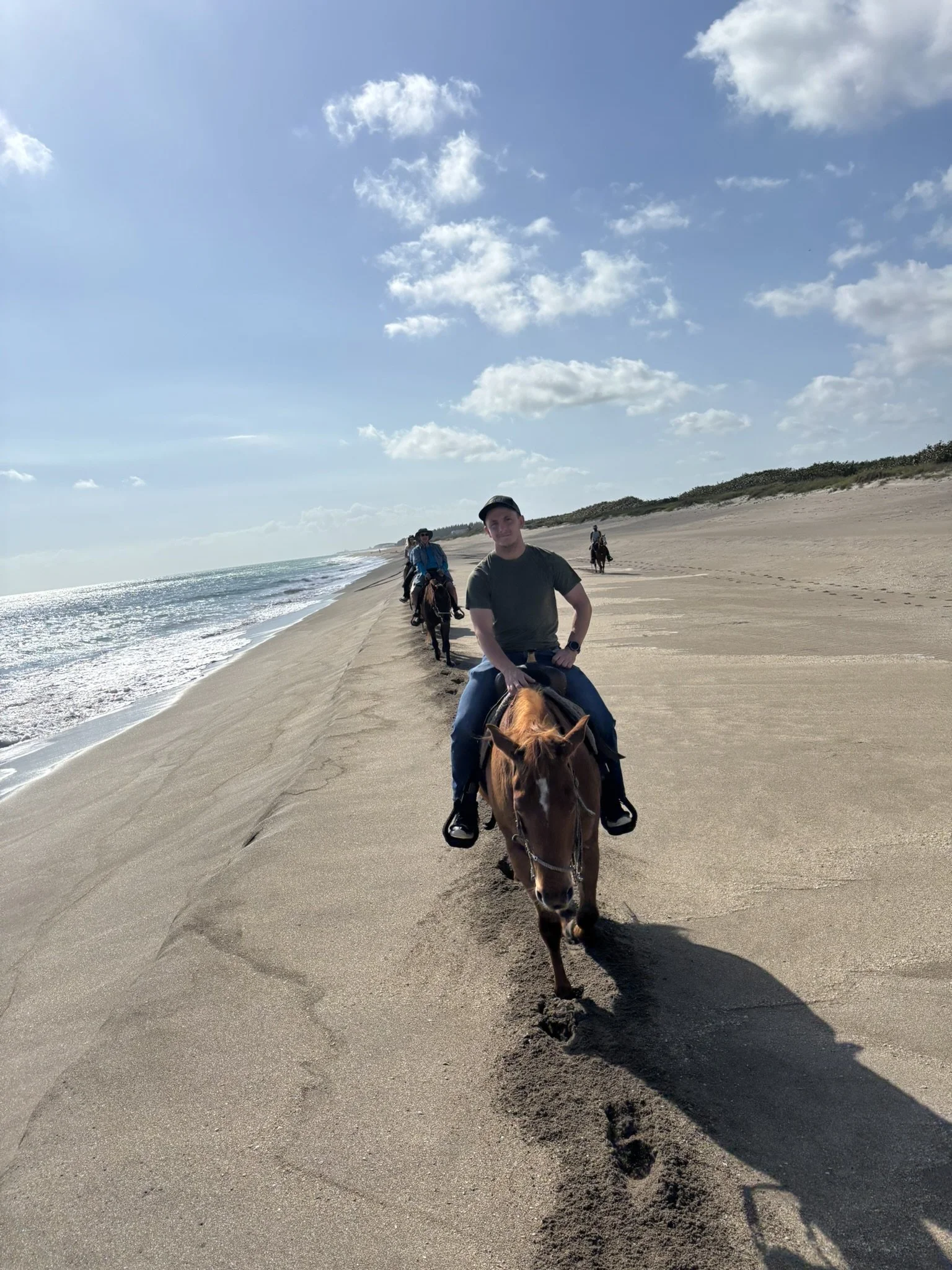 People riding horses along a sandy beach on a sunny day with partly cloudy sky.
