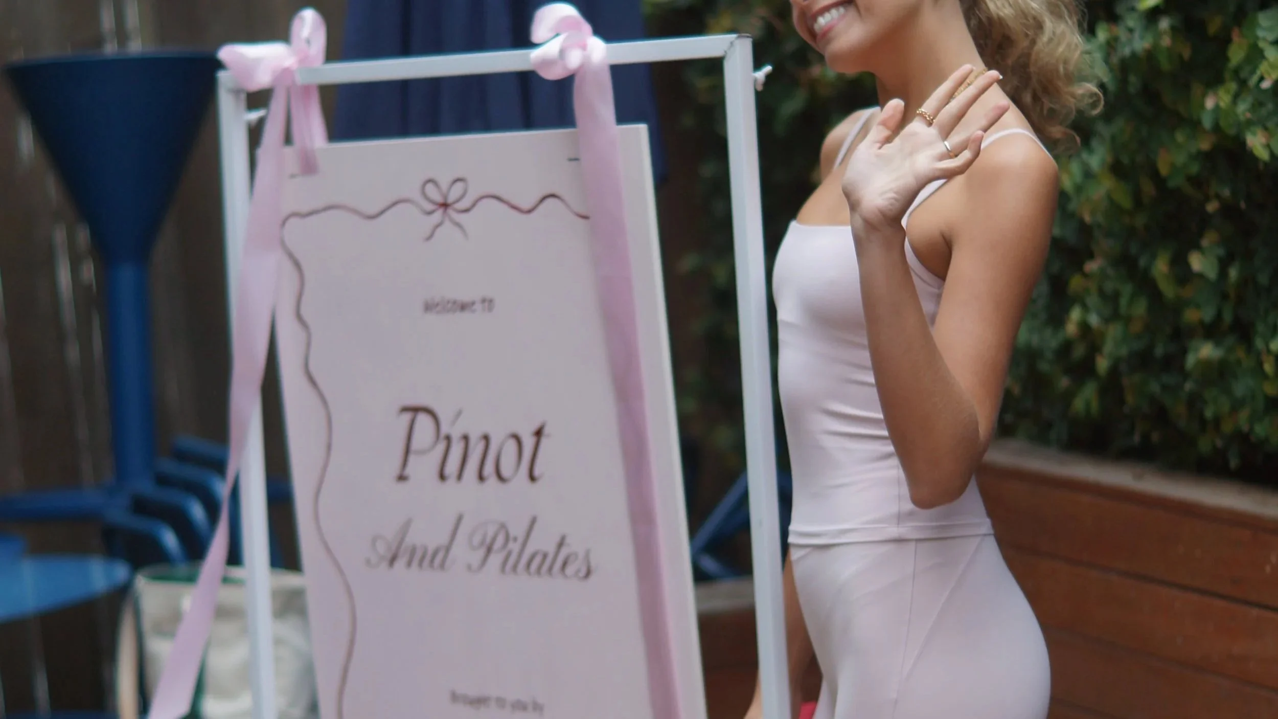 A woman in a white dress waving near a sign for Pinot and Pilates, with a blurred outdoor background.