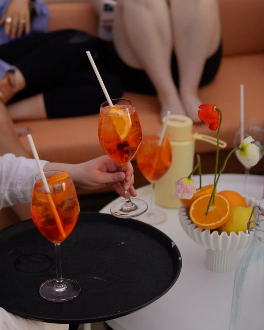 Three glasses of orange-colored beverages with straws on a table, alongside a white bowl of sliced oranges and flowers, with people sitting in the background.