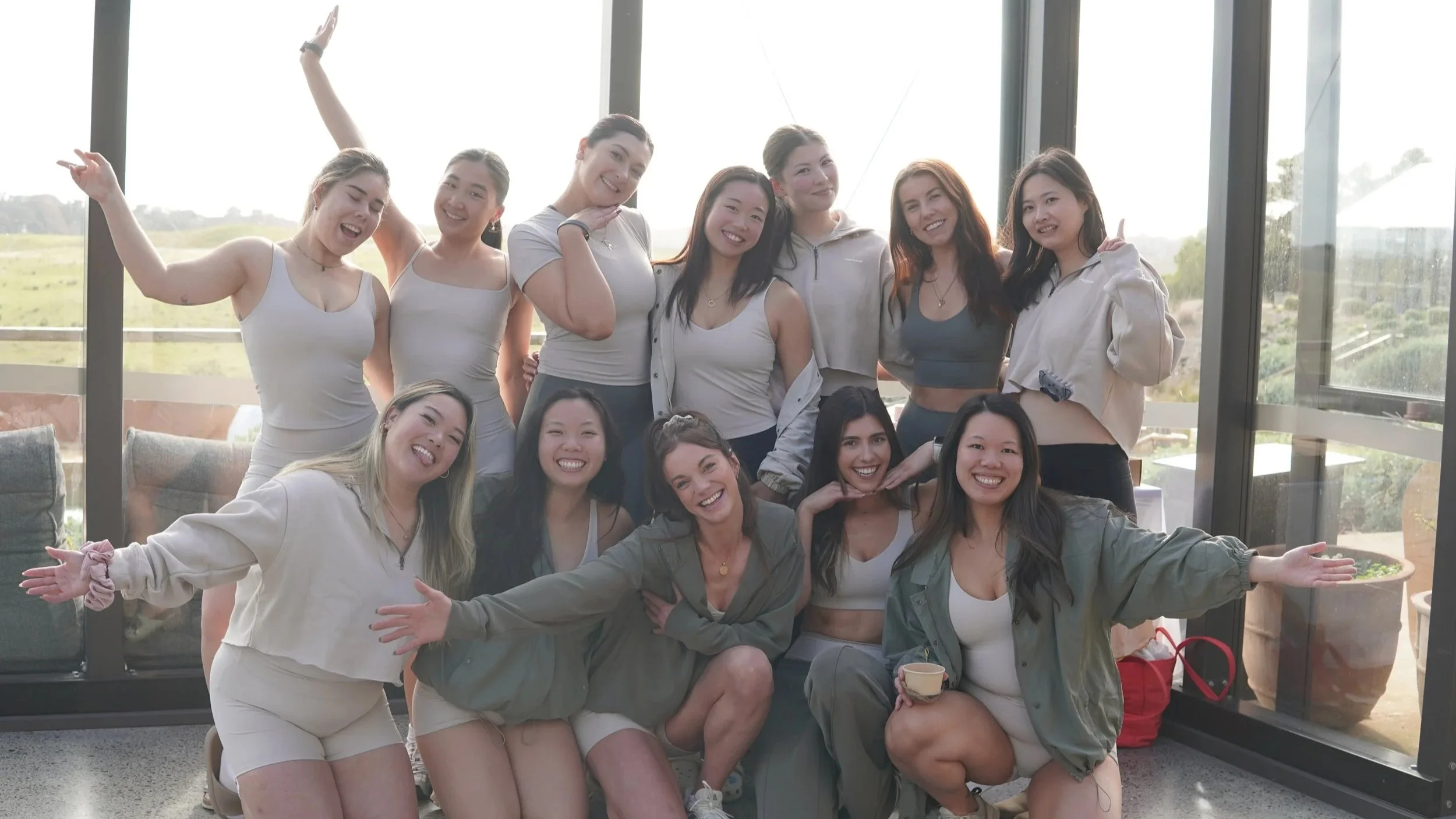 A group of fifteen women posing inside a sunlit room with floor-to-ceiling windows, outdoors scenery visible, most wearing casual workout clothes or dresses, smiling and making playful gestures.