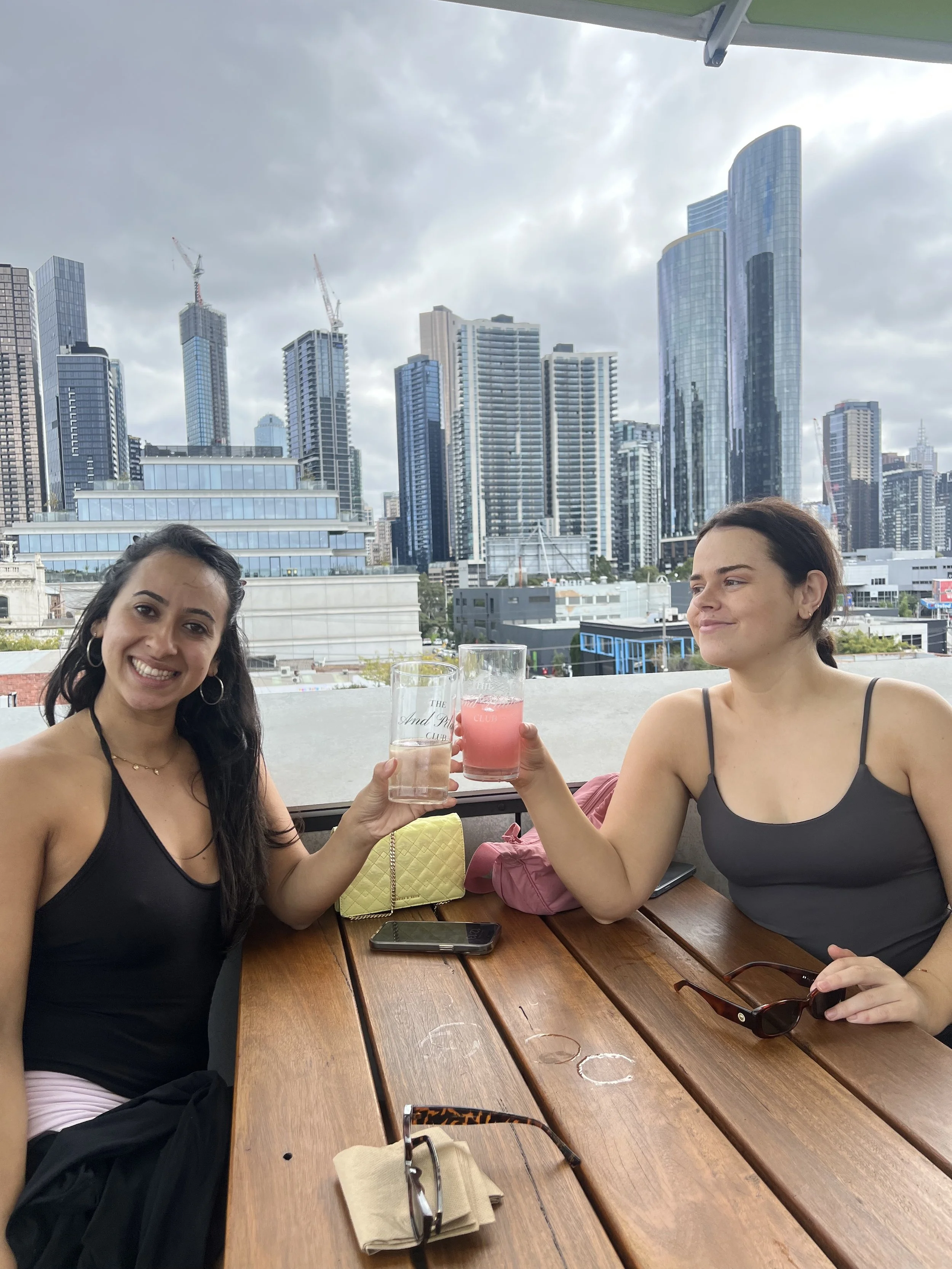 Two women sitting at a wooden table outdoors in a city with tall skyscrapers in the background, raising their glasses in a toast and smiling.