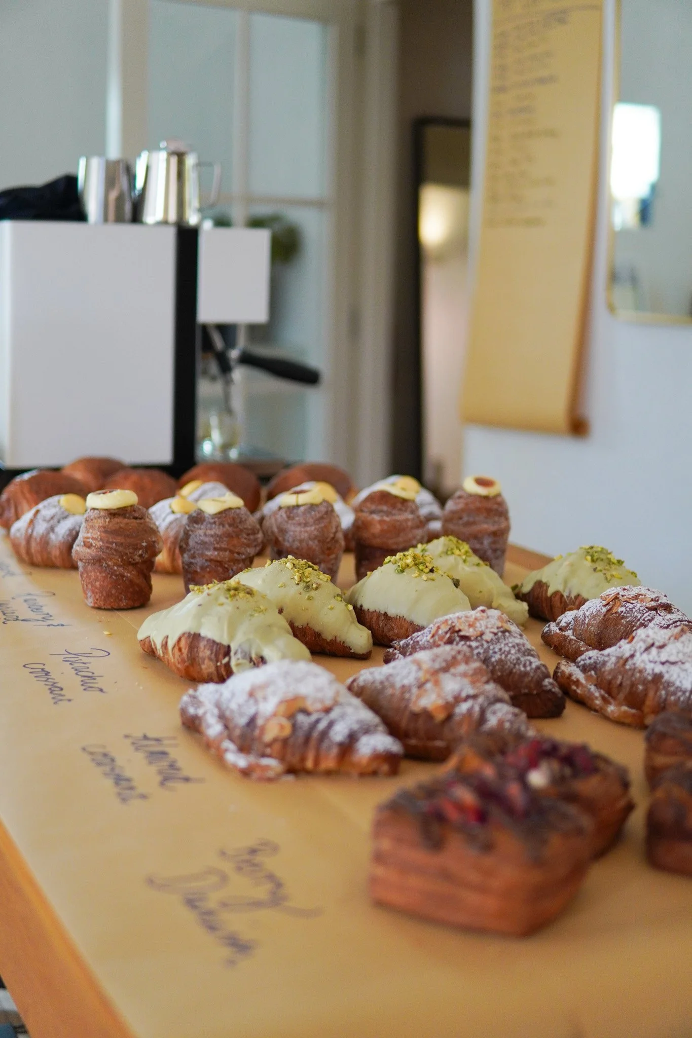Assorted pastries, including croissants with green icing, pastries topped with white chocolate and pistachios, and others dusted with powdered sugar, arranged on a table in a cafe or bakery.