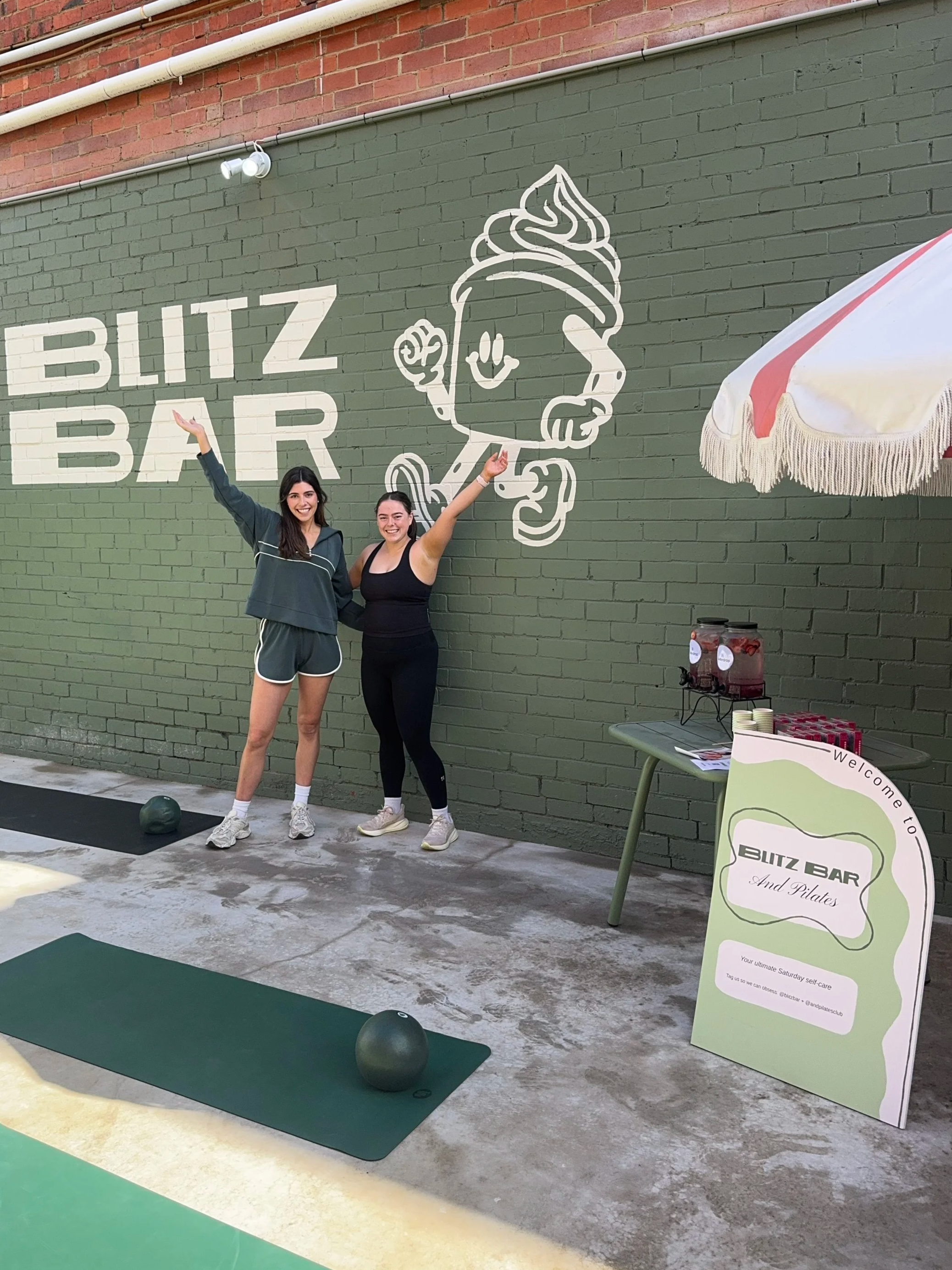Two women smiling and posing with raised arms in front of a green brick wall with a white cartoon ice cream cone mascot and the words 'Blitz Bar'. There are workout mats and kettlebells on the ground, and a table with a drink station and a sign welcoming to Blitz Bar and Pilates.
