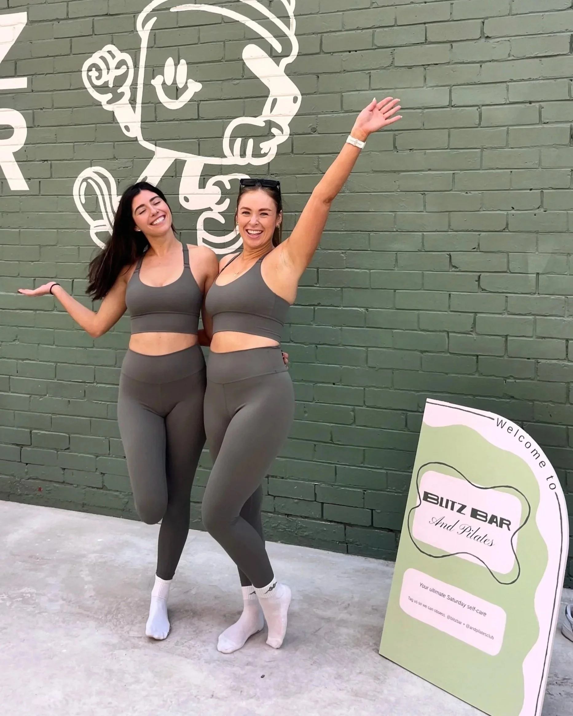 Two women wearing matching athletic outfits posing cheerfully in front of a green brick wall with a chalk outline of a cartoon character. A sign on the ground reads 'Blitz Bar and Pilates'.