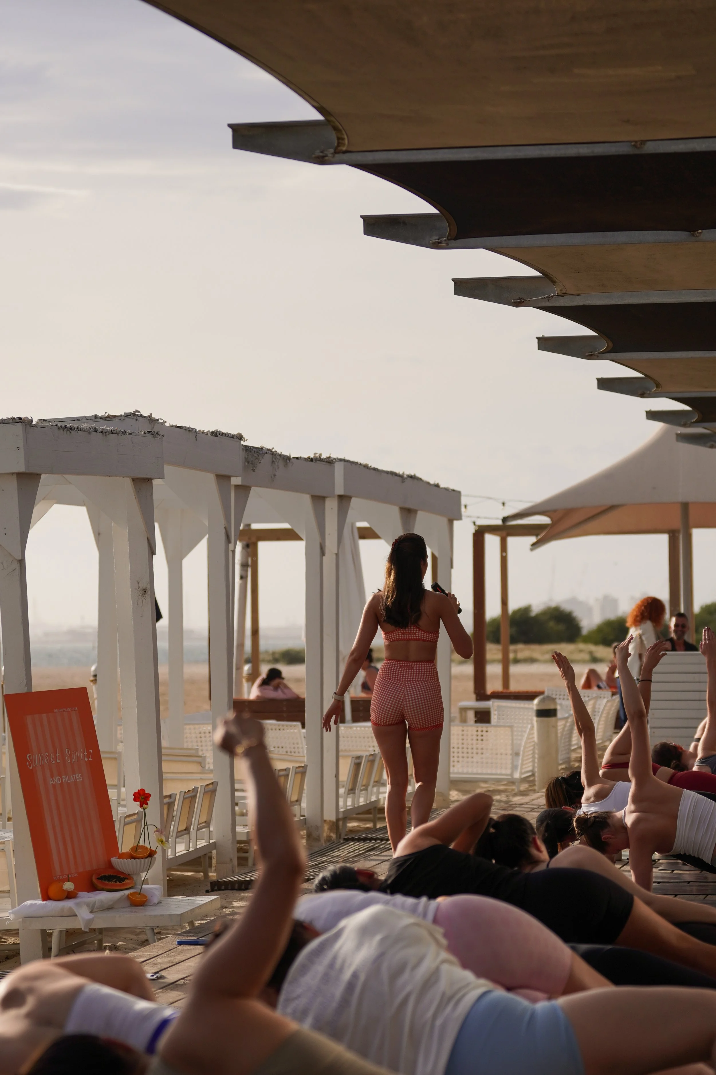 People participating in a yoga class on a beach during sunset, with a woman leading the session facing the group, some participants doing poses.