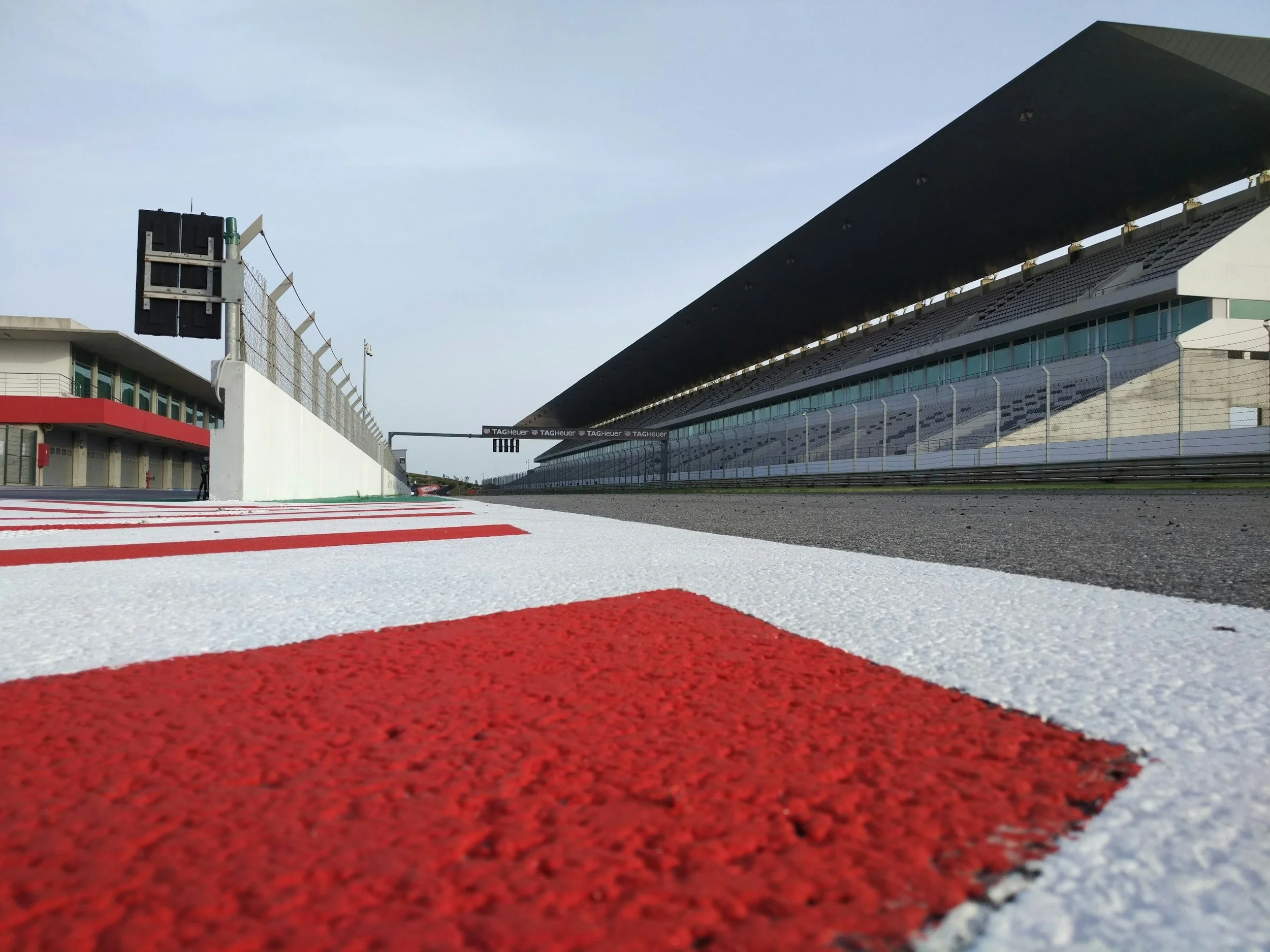 Empty race track with red and white curbing, grandstand in the background, and clear sky.