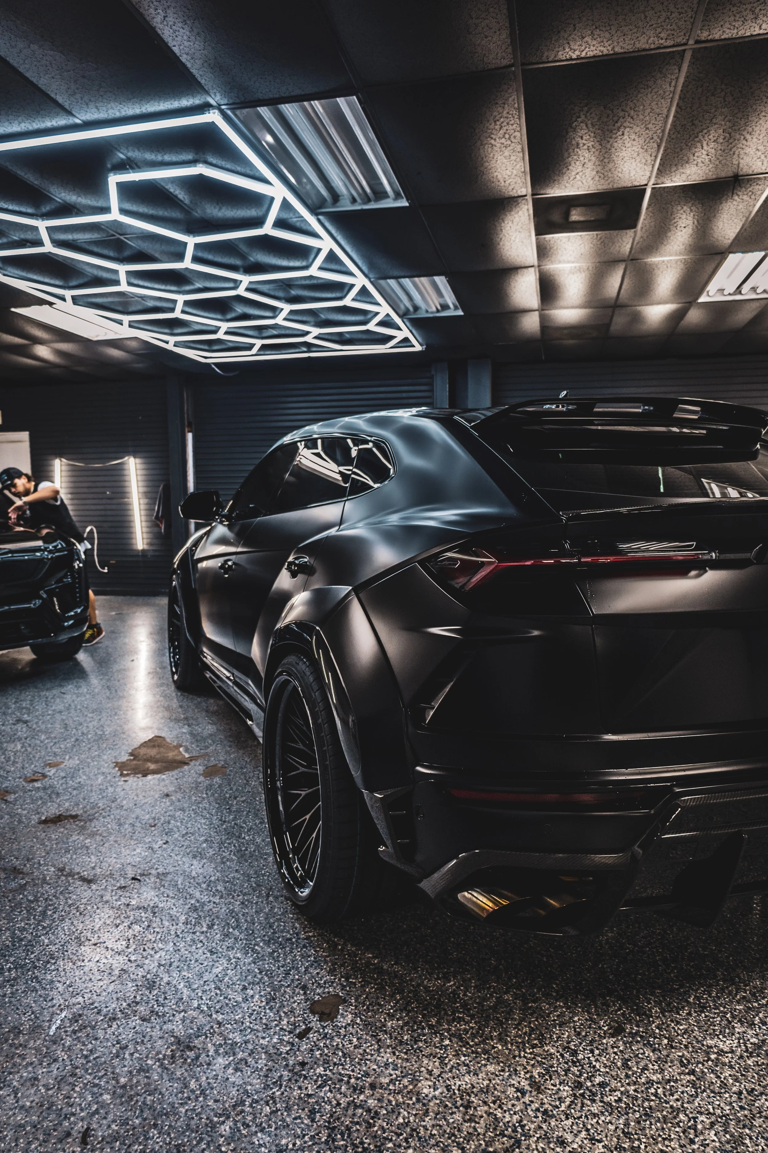 A sleek, black, modern car parked indoors with a person inspecting another car in the background, and contemporary geometric ceiling lights.