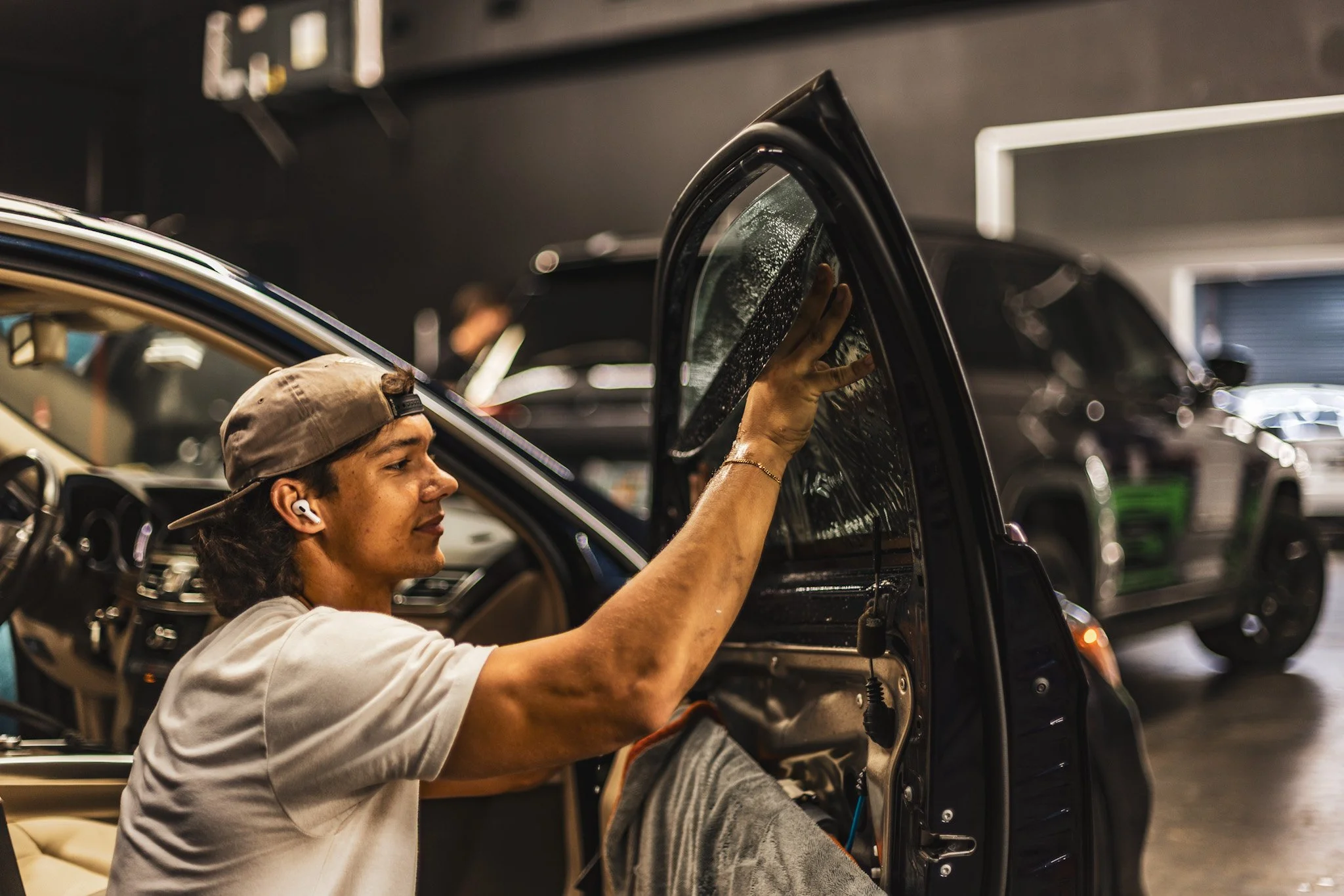 A young male mechanic wearing a cap and earbud working on a car inside a garage, with other vehicles visible in the background.