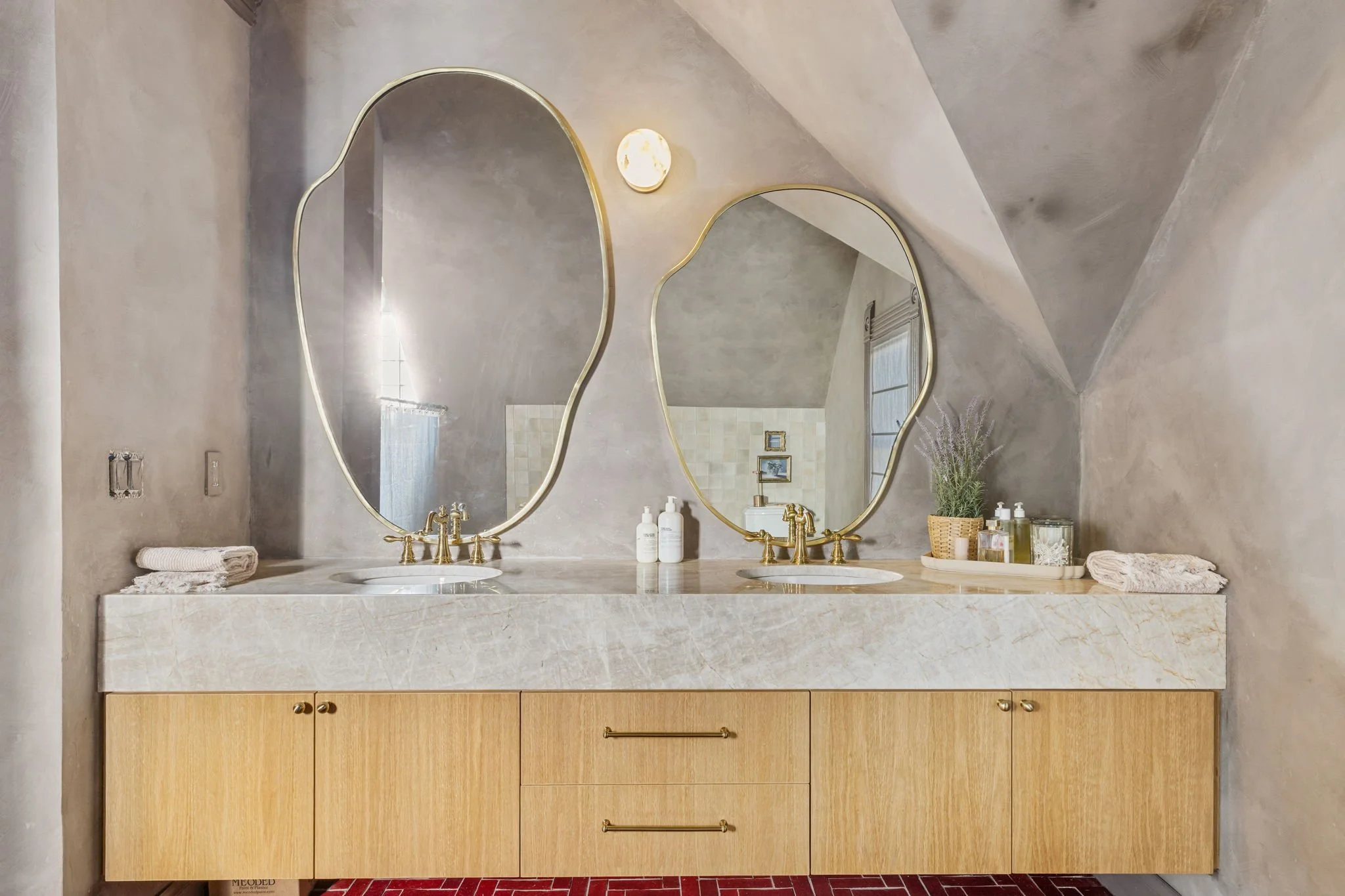 A bathroom vanity with two gold-framed mirrors, gold faucets, and a marble countertop. The vanity has a wooden cabinet with drawers and gold handles. There are two soap dispensers, a small tray with toiletries, a potted plant, and towels on the count