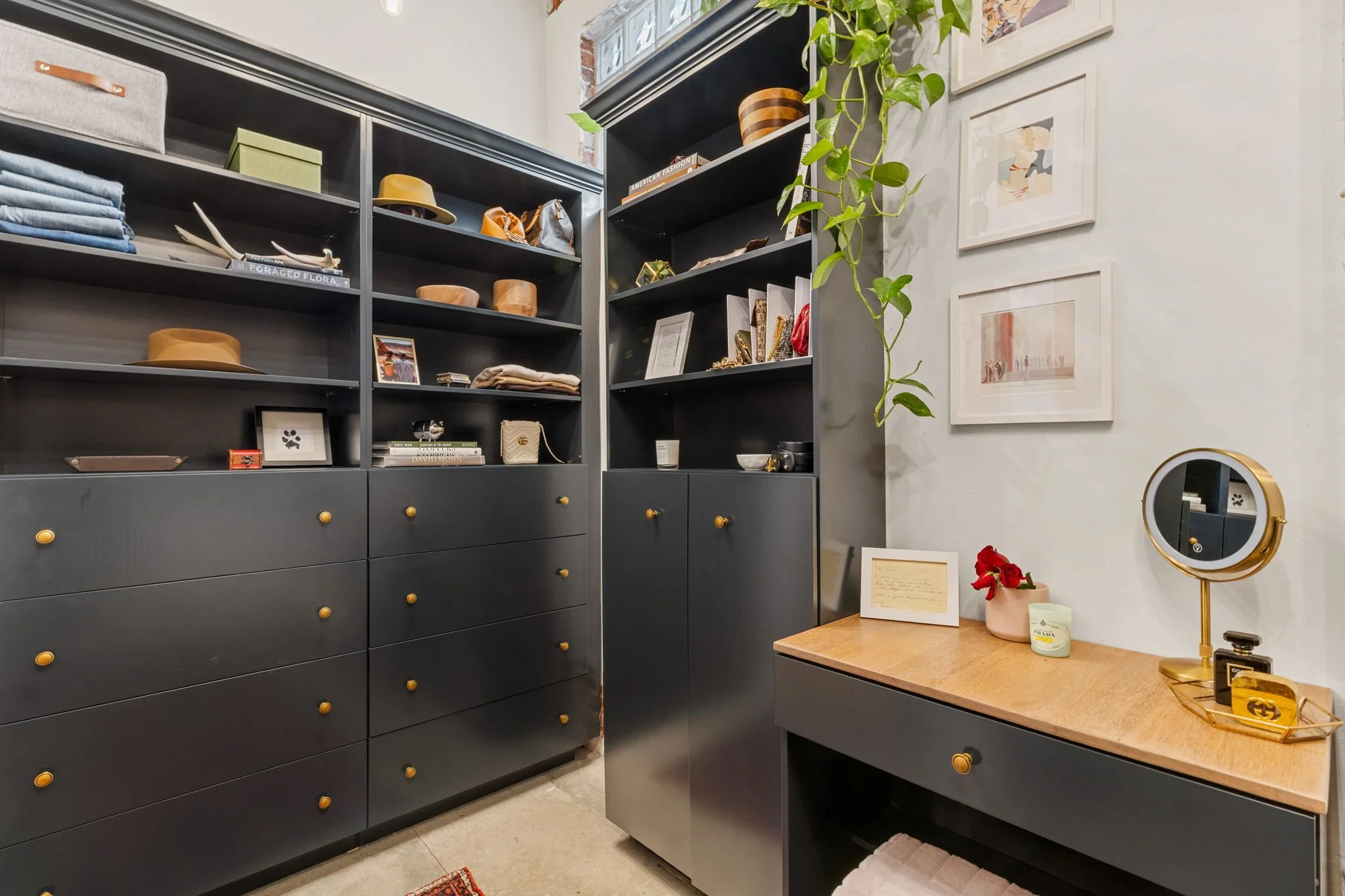 A walk-in closet with black shelves and drawers filled with hats, books, and accessories. A small wooden table with a mirror, candles, and framed photos is on the right, and a hanging plant drapes down the corner.