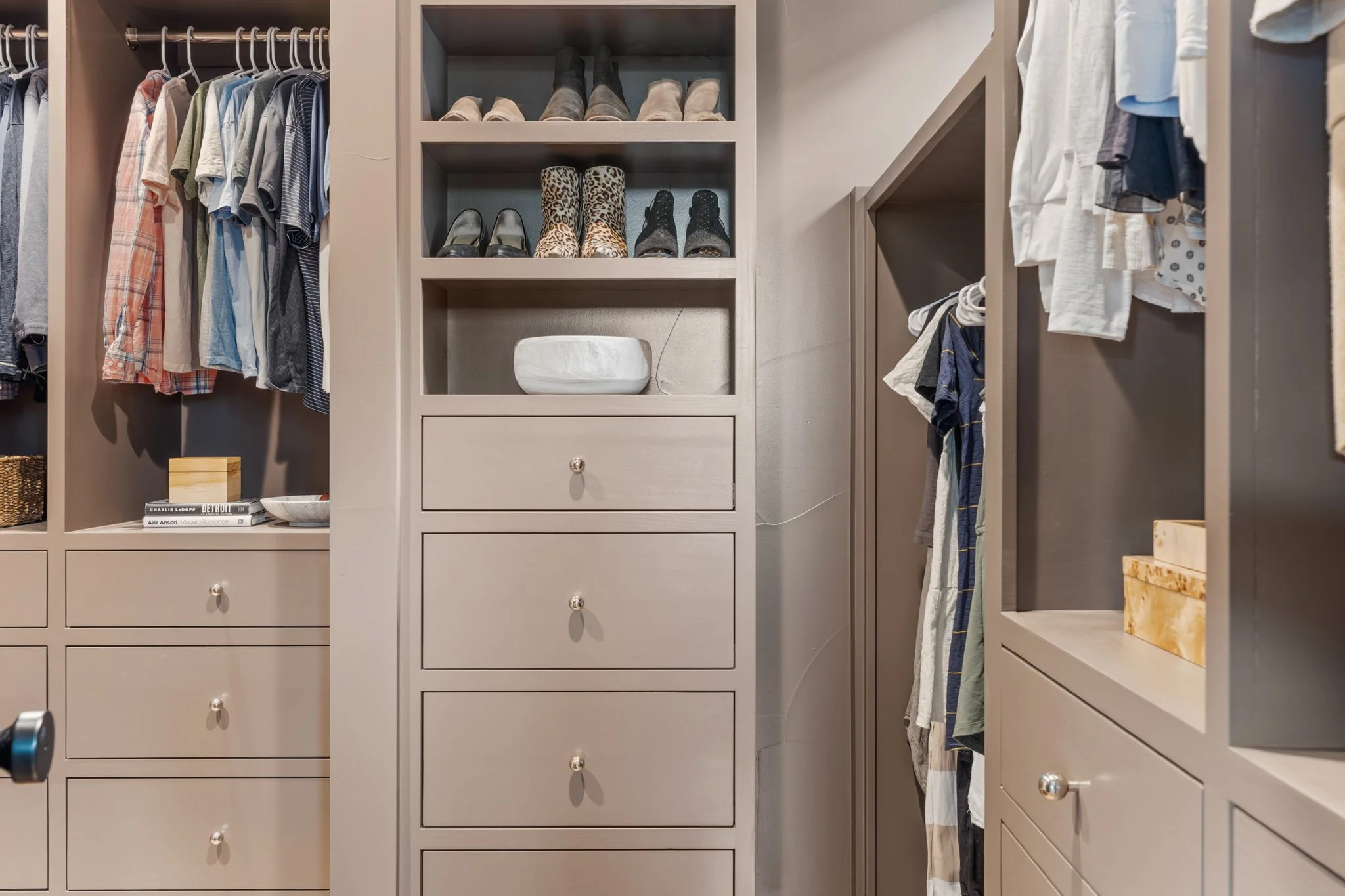 A modern walk-in closet with open shelves and hanging space filled with folded clothes, shirts, and shoes. There's a drawer section and various boxes and decorative items on the shelves.
