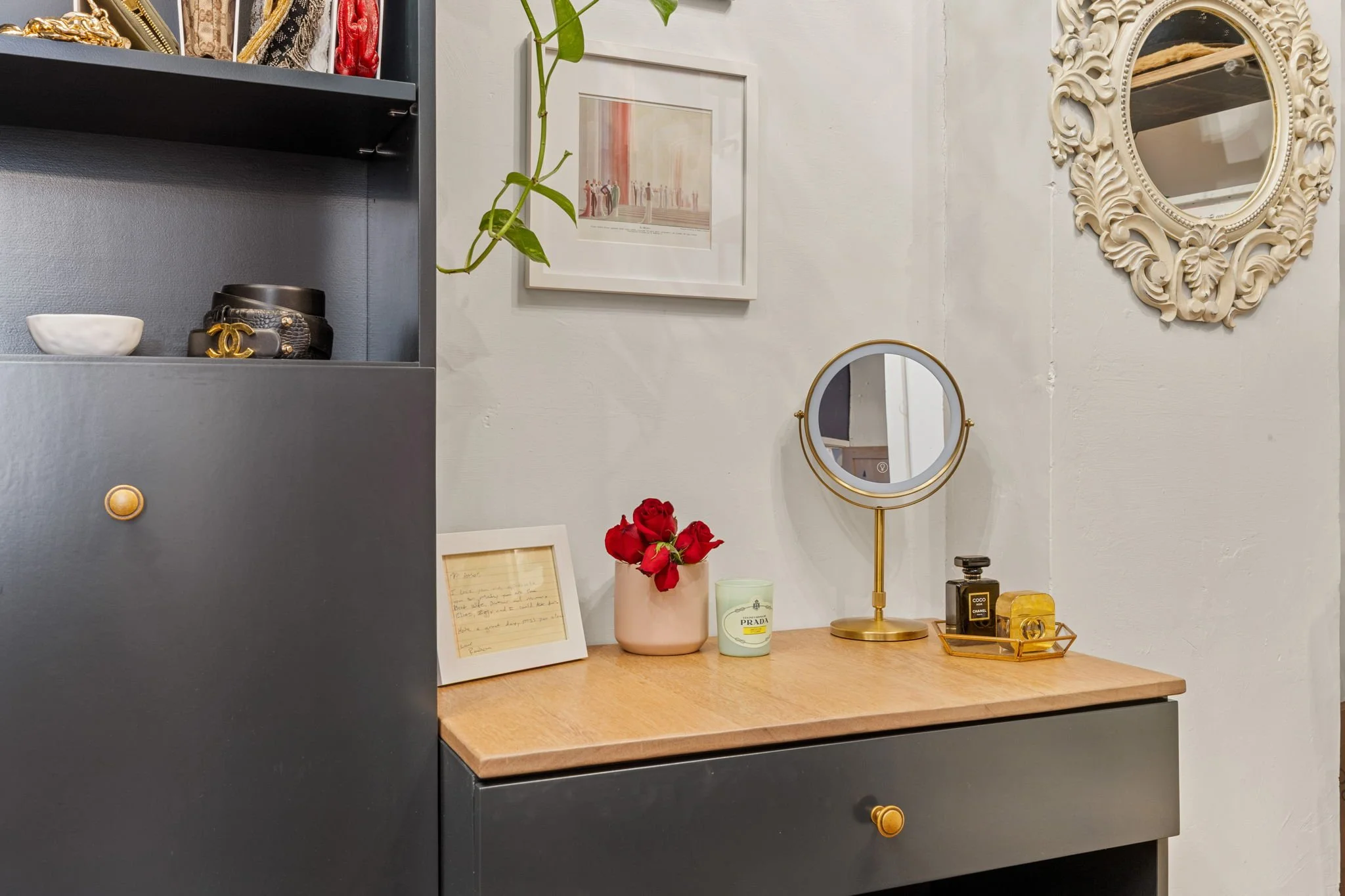 A vanity or side table with a pink vase of red roses, a small white picture frame, a scented candle, and a small round mirror in a gold stand. There are decorative framed pictures and a large ornate mirror on the wall.
