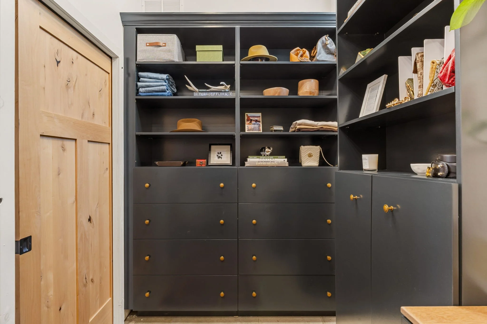 A black shelving unit with several shelves and drawers, containing hats, books, framed photos, handbags, and decorative items, situated in a room with a wooden door on the left.