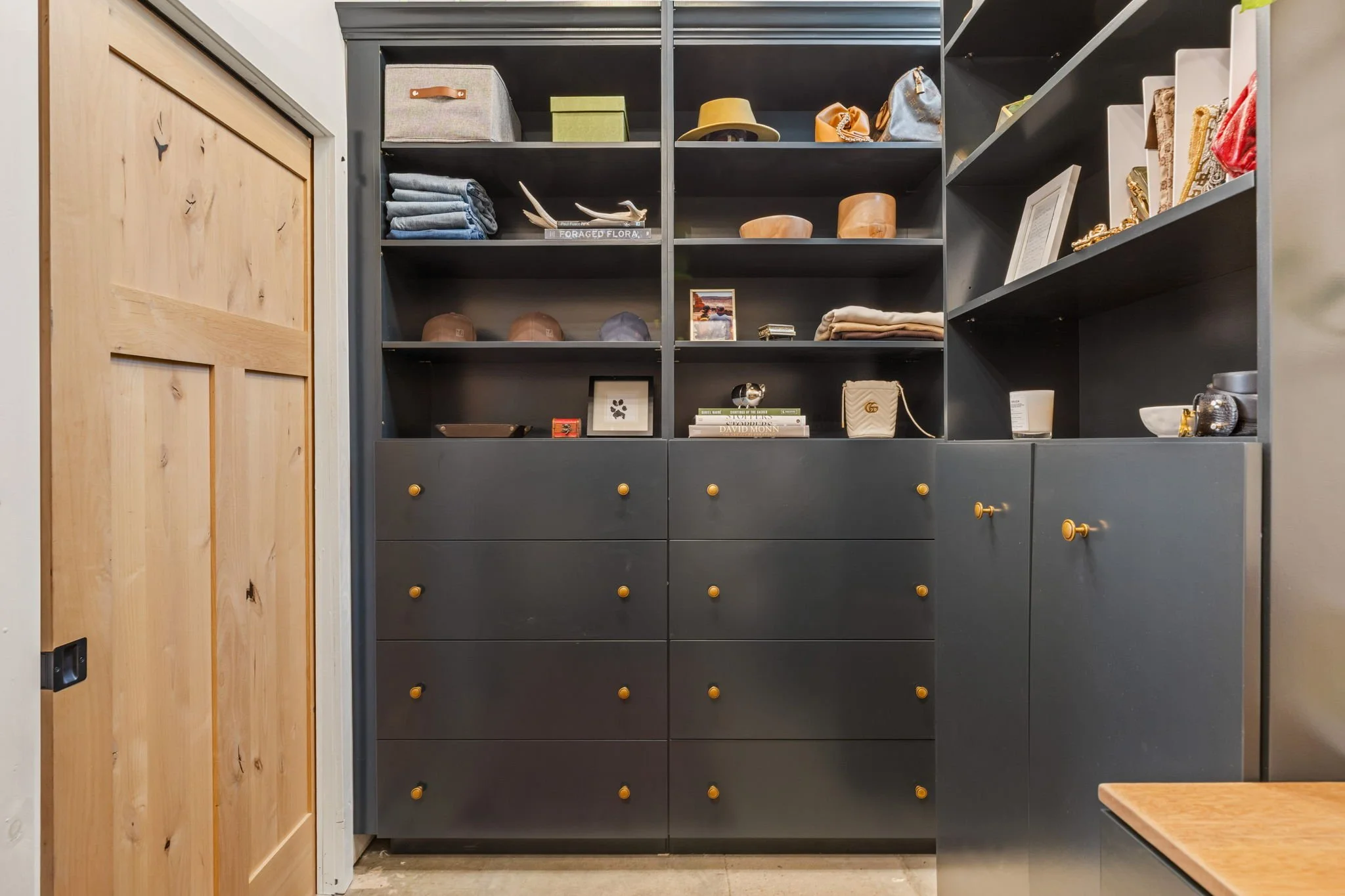 Dark gray shelving unit with multiple open shelves and drawers, decorated with hats, books, picture frames, folded clothes, and decorative objects, next to a wooden door.