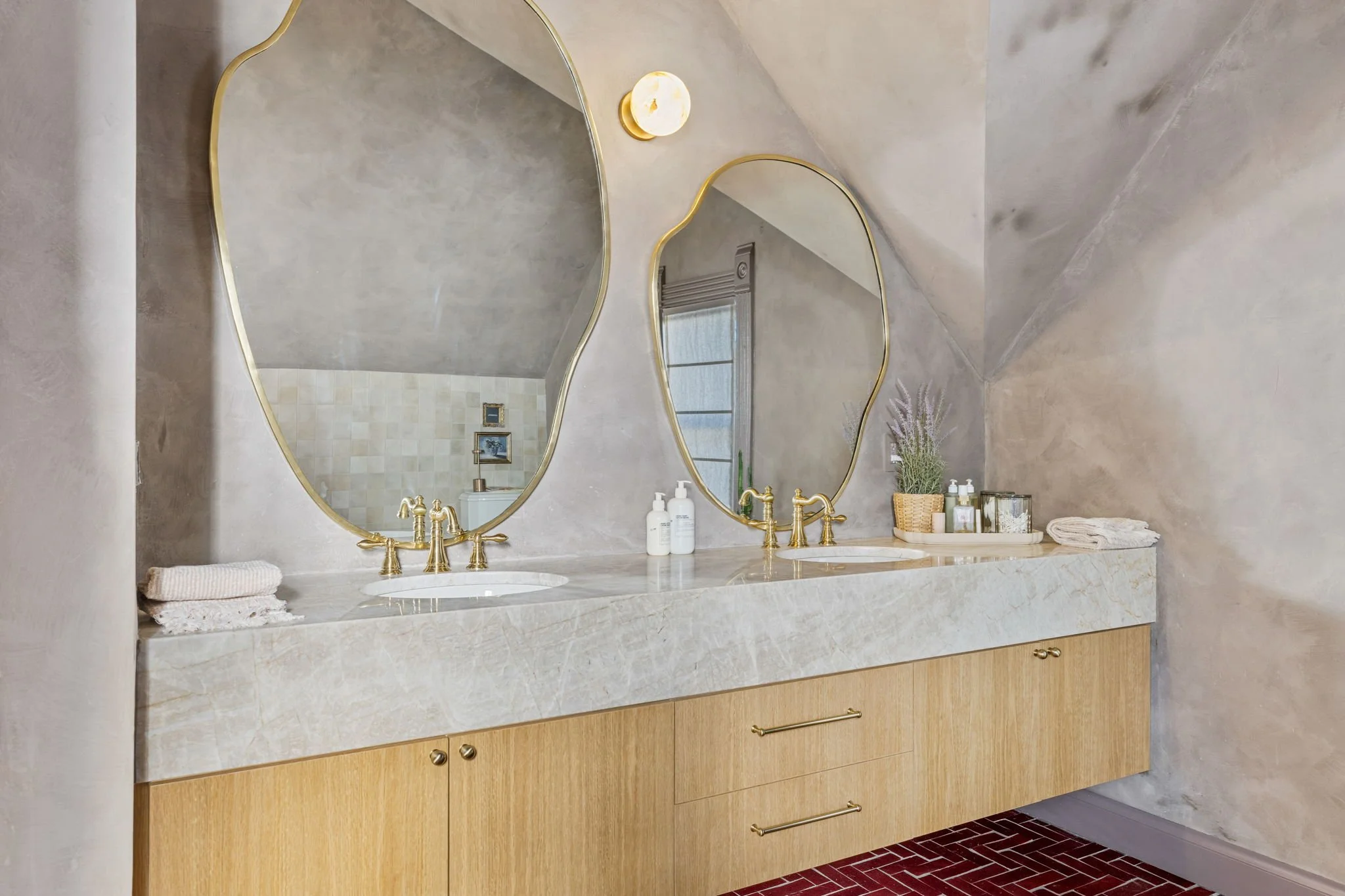 A bathroom vanity with a marble countertop, two mirrors with gold frames, gold fixtures, and decorative items including towels, soap dispensers, and a potted plant.