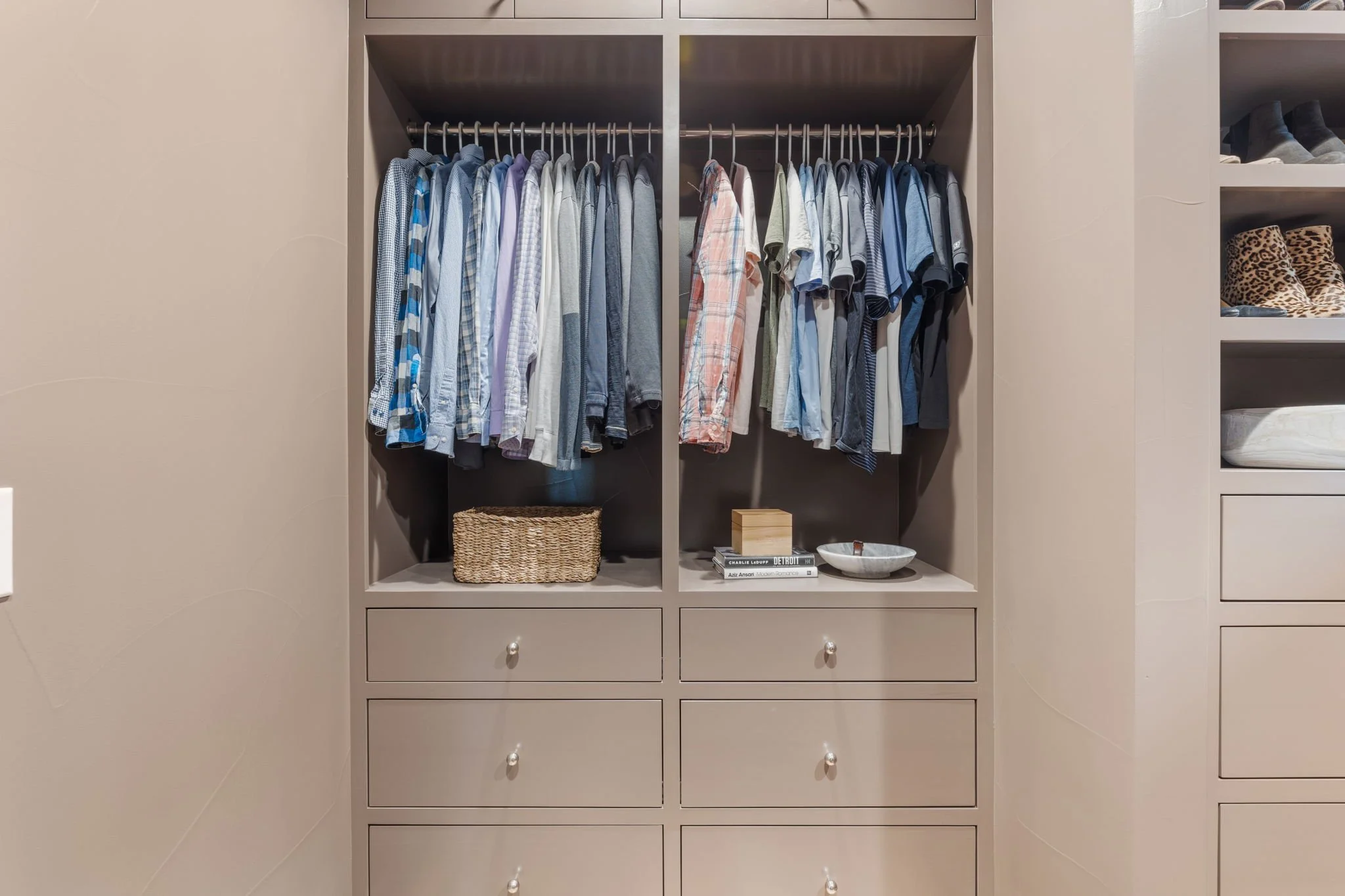 Open closet with hanging shirts on two rods, a woven basket, a stack of books, a small wooden box, and a white bowl inside, surrounded by beige walls and cabinets.