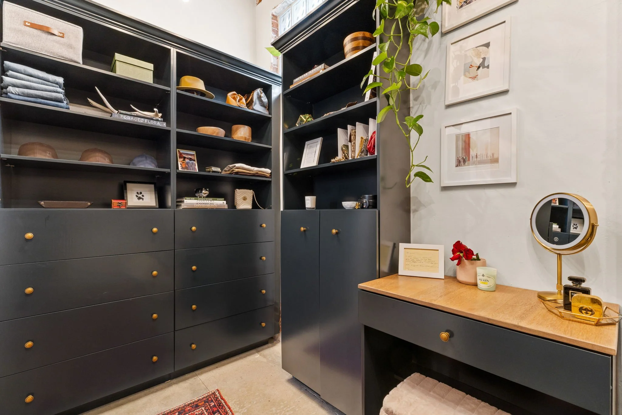A walk-in closet with black cabinets filled with clothes, hats, and accessories. A light-colored wall with framed artwork, a small plant, and a wooden dresser with decorative items, including a mirror, a candle, and a book.
